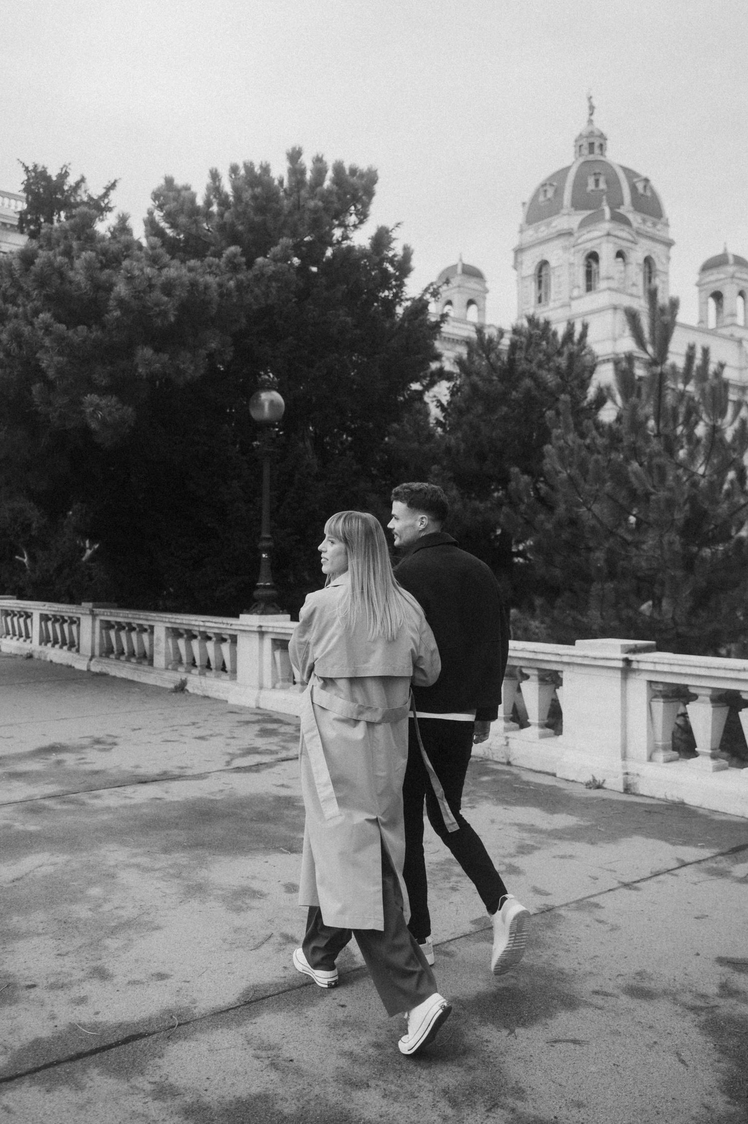 A couple walks together on a path with the domed Kunsthistorisches Museum in Vienna visible in the background.