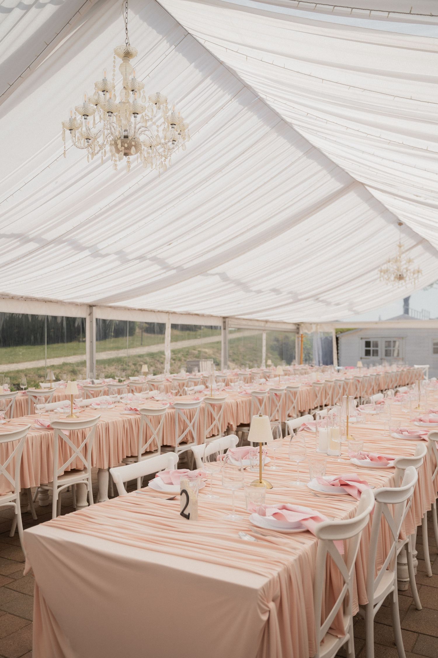 Elegant wedding reception tent with pink striped tablecloths, crystal chandeliers, and white cross-back chairs.