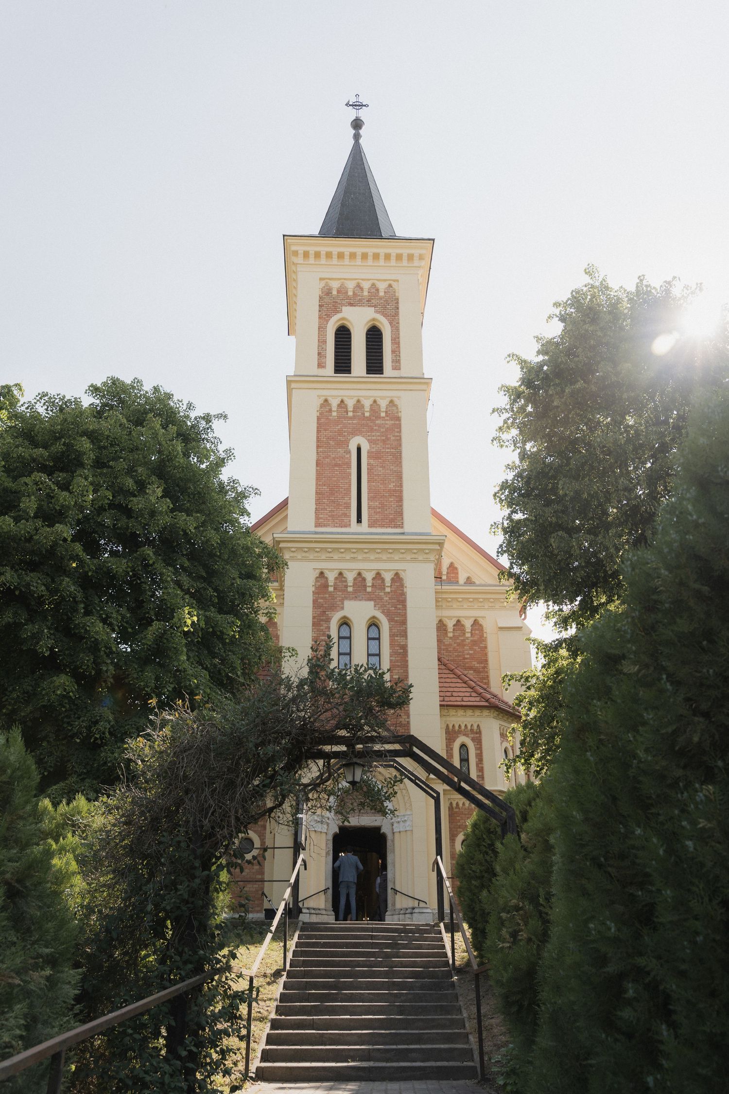 Historic church with tall bell tower framed by lush green trees and stone staircase entrance.