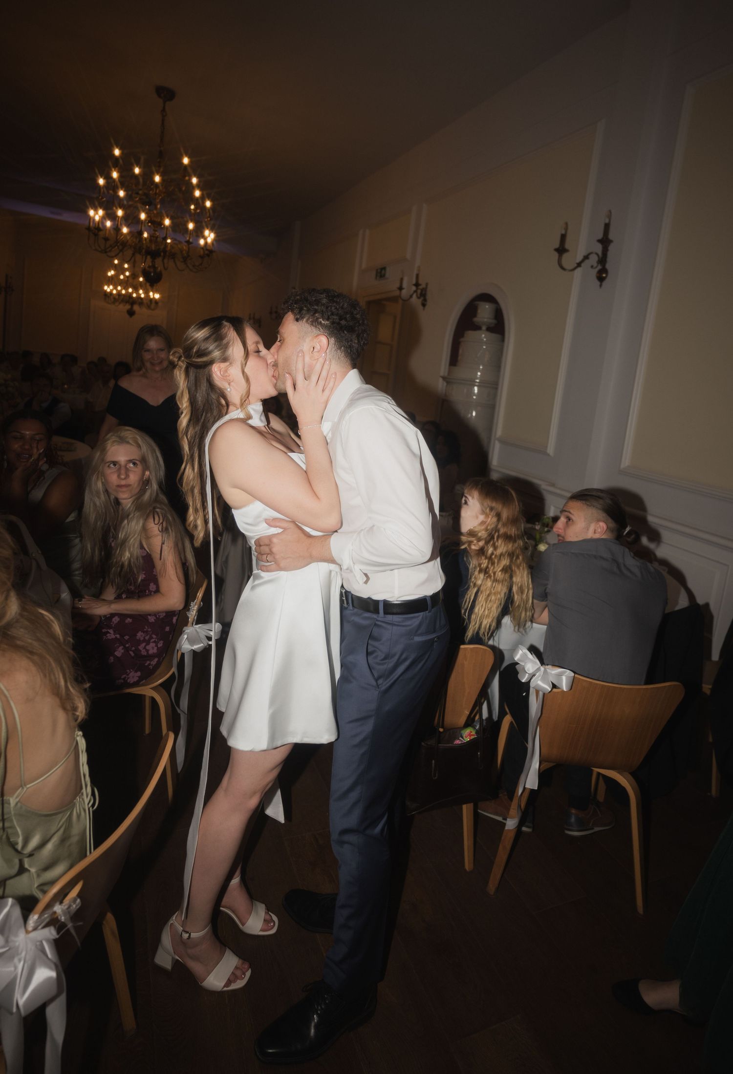 Couple sharing romantic kiss during wedding reception while guests watch in candlelit elegant venue.