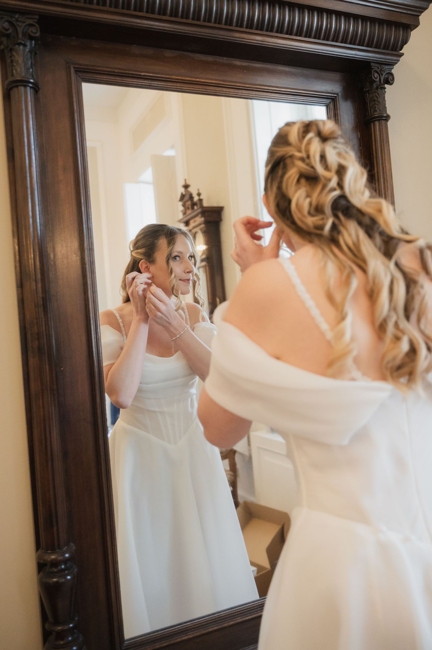 Bride in white off-shoulder gown adjusting earring while looking into ornate wooden mirror.