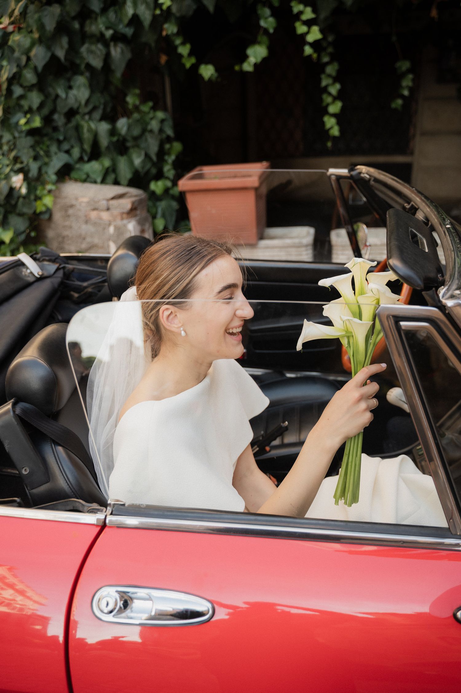 Bride holding white calla lily bouquet sits in red vintage convertible car.