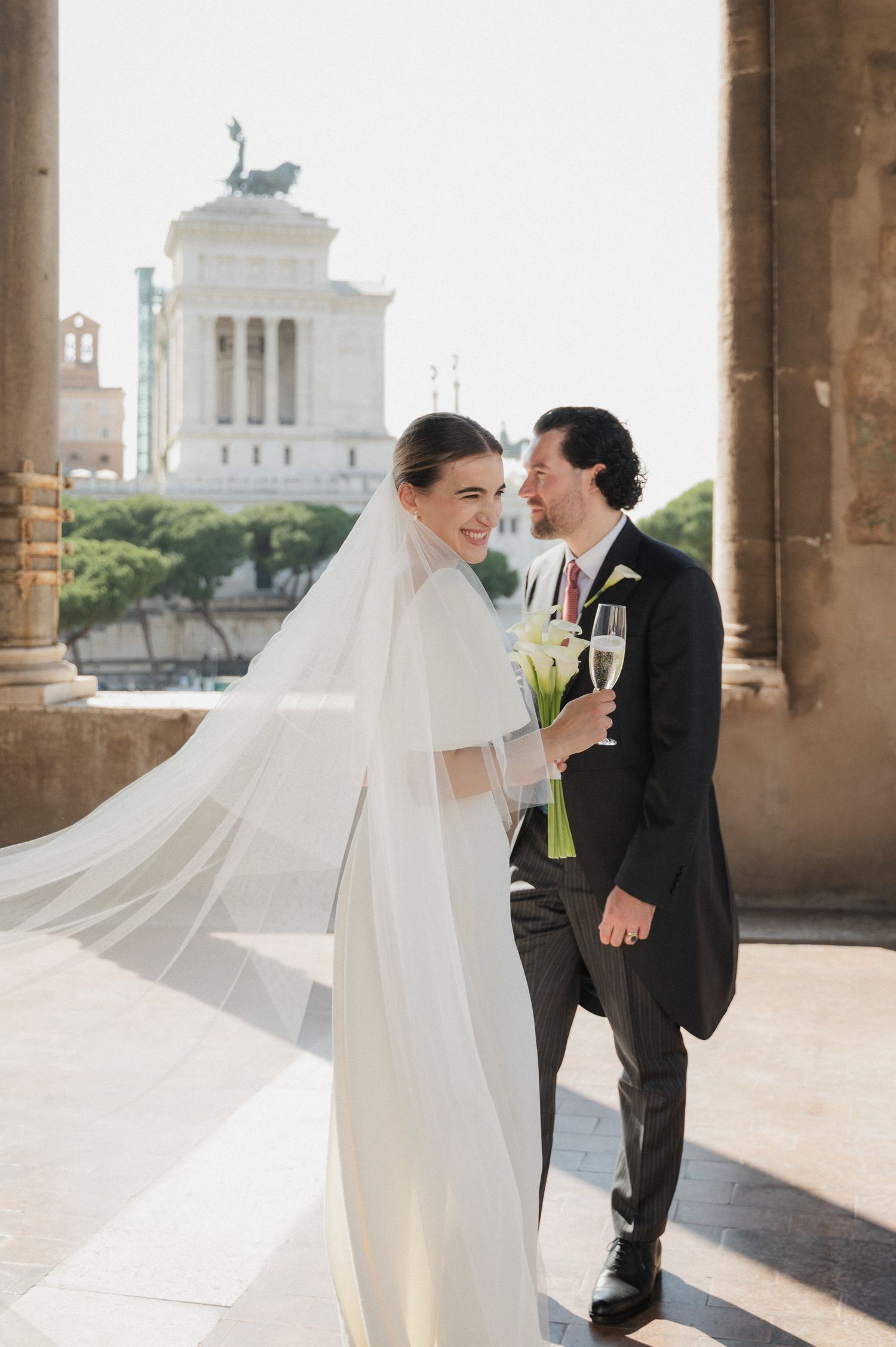 A wedding couple shares a romantic moment under stone archways with a historic building visible in the background.