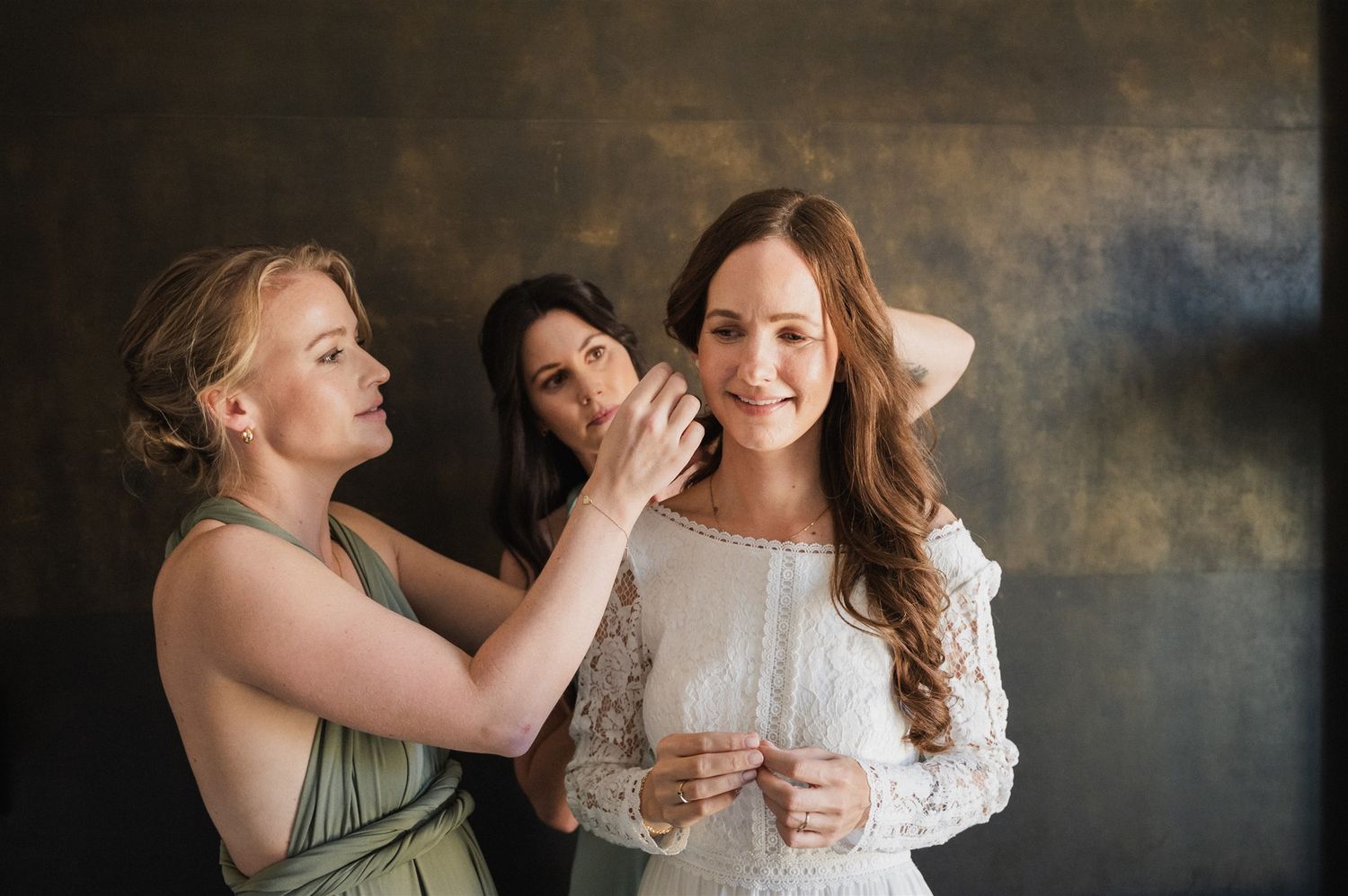 Bridesmaids in sage green dresses share a candid moment against a dark backdrop.