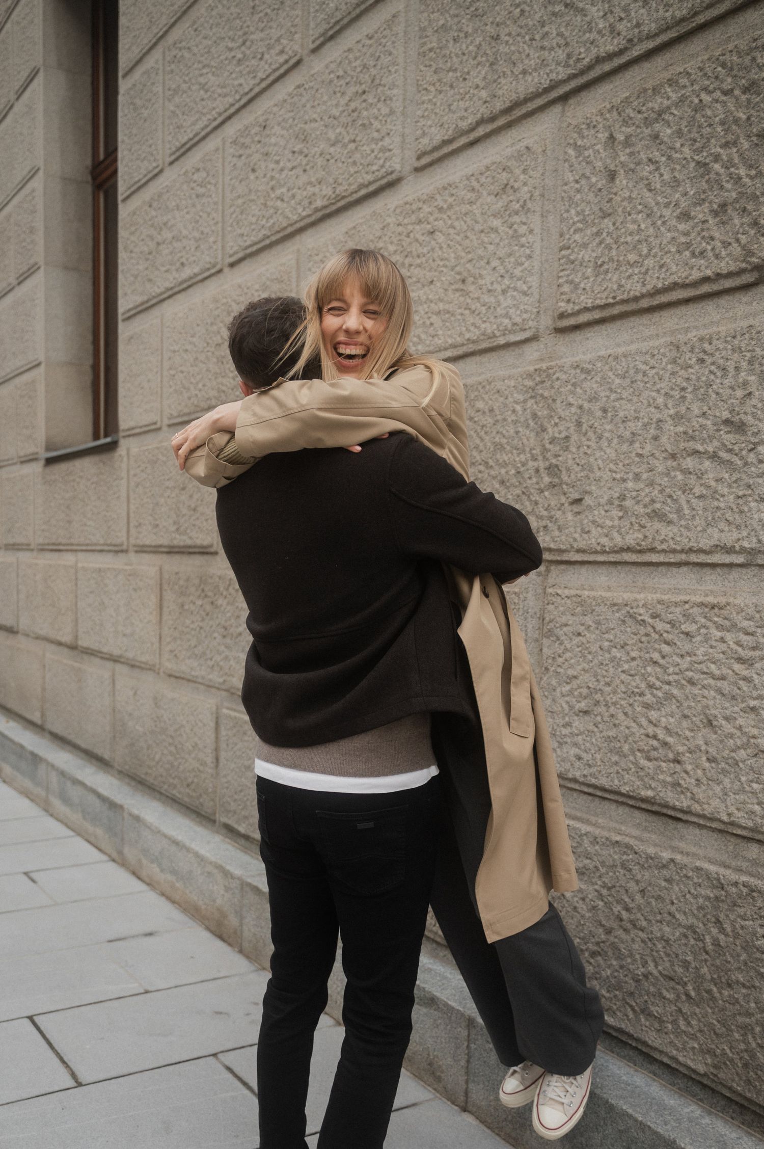 Two people embrace in a warm hug against a textured stone wall while wearing casual attire.