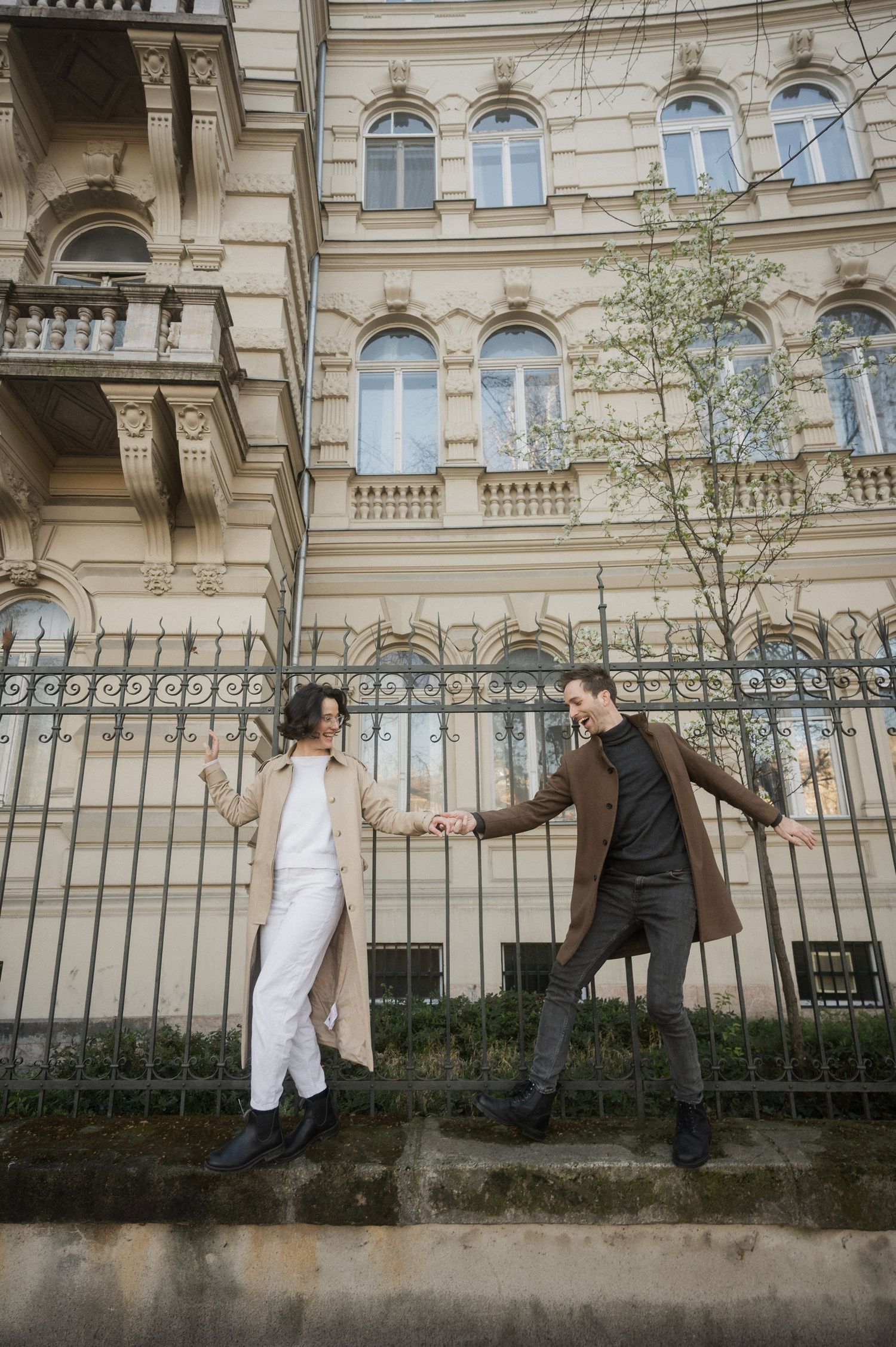 A couple poses playfully outside an ornate historic building with arched windows and iron fencing in an urban setting.