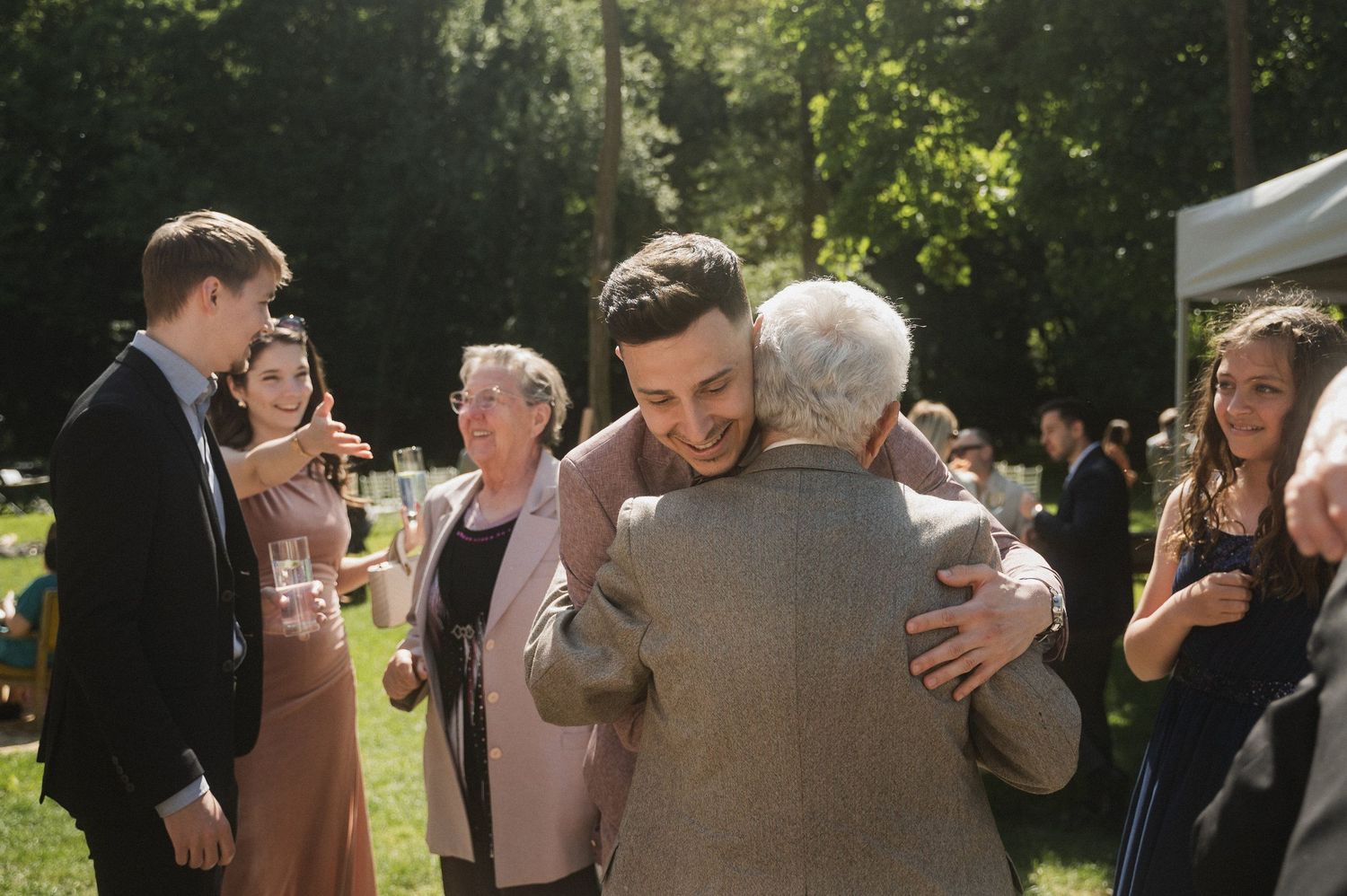 People gather at an outdoor event in a sunny garden setting sharing warm embraces and conversations.