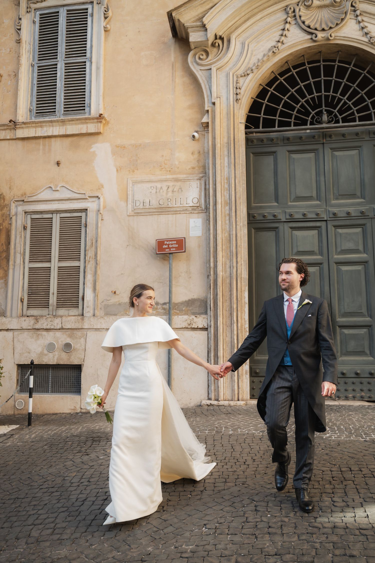 A couple in wedding attire playfully pose on a cobblestone street in front of vintage European building doors.