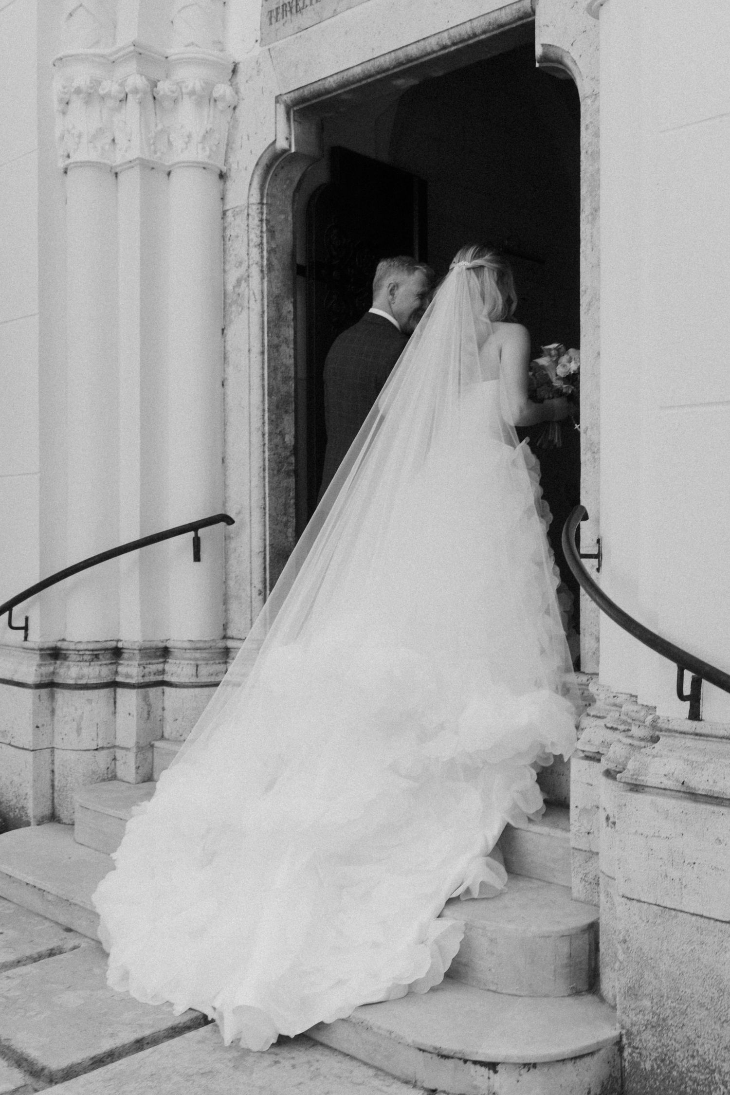 Bride in flowing gown and cathedral veil enters church with groom on wedding day, black and white photo.