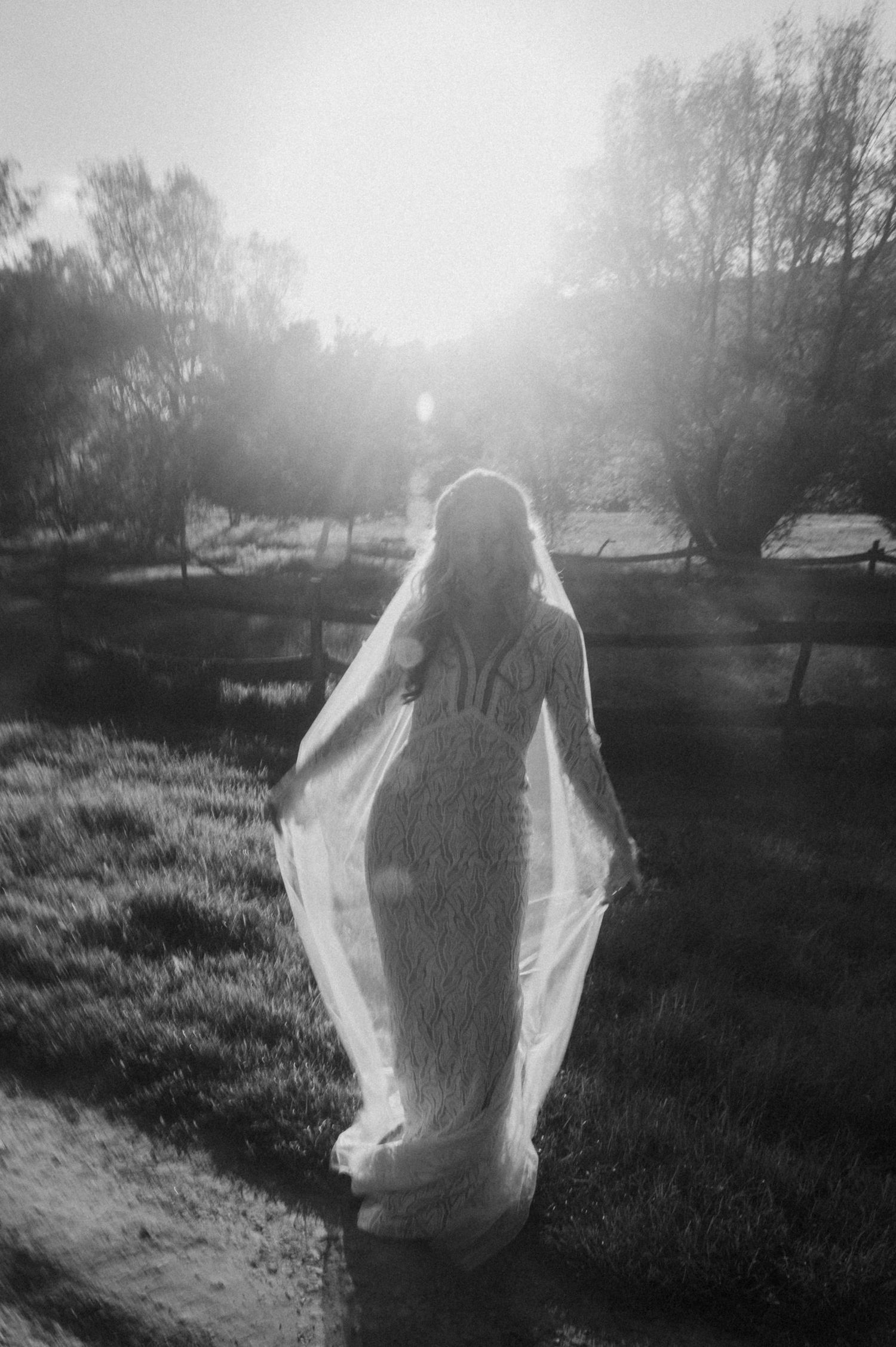 A ghostly figure in white clothing stands backlit in a misty field creating an ethereal black and white scene.