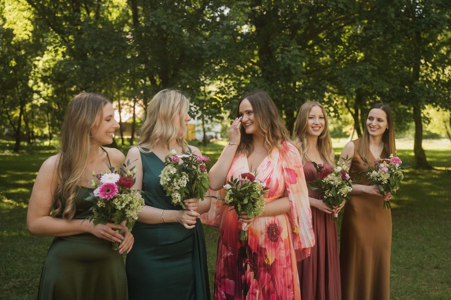 A group of bridesmaids in coral and brown dresses stand together holding colorful bouquets in an outdoor garden setting.