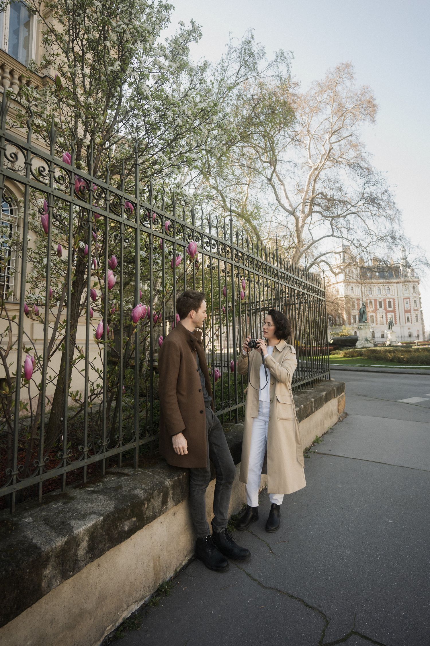 Two people in coats have a conversation while standing along a wrought iron fence on a city sidewalk.