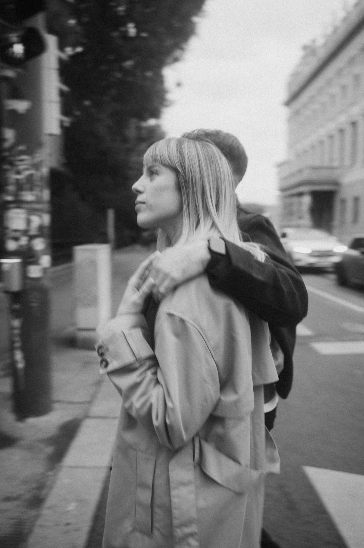 A black and white photo shows a person in a light trench coat walking on a city street.