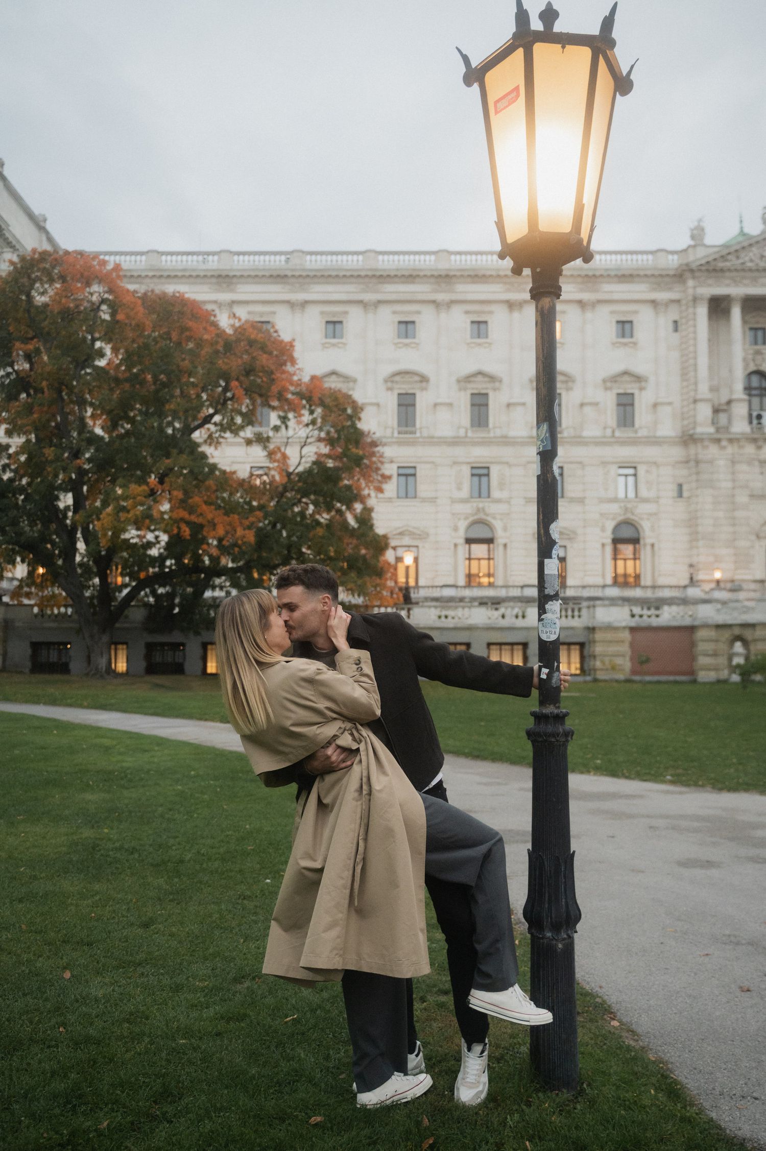 A person in a beige coat poses near a historic white building with autumn trees and a vintage lamppost in view.