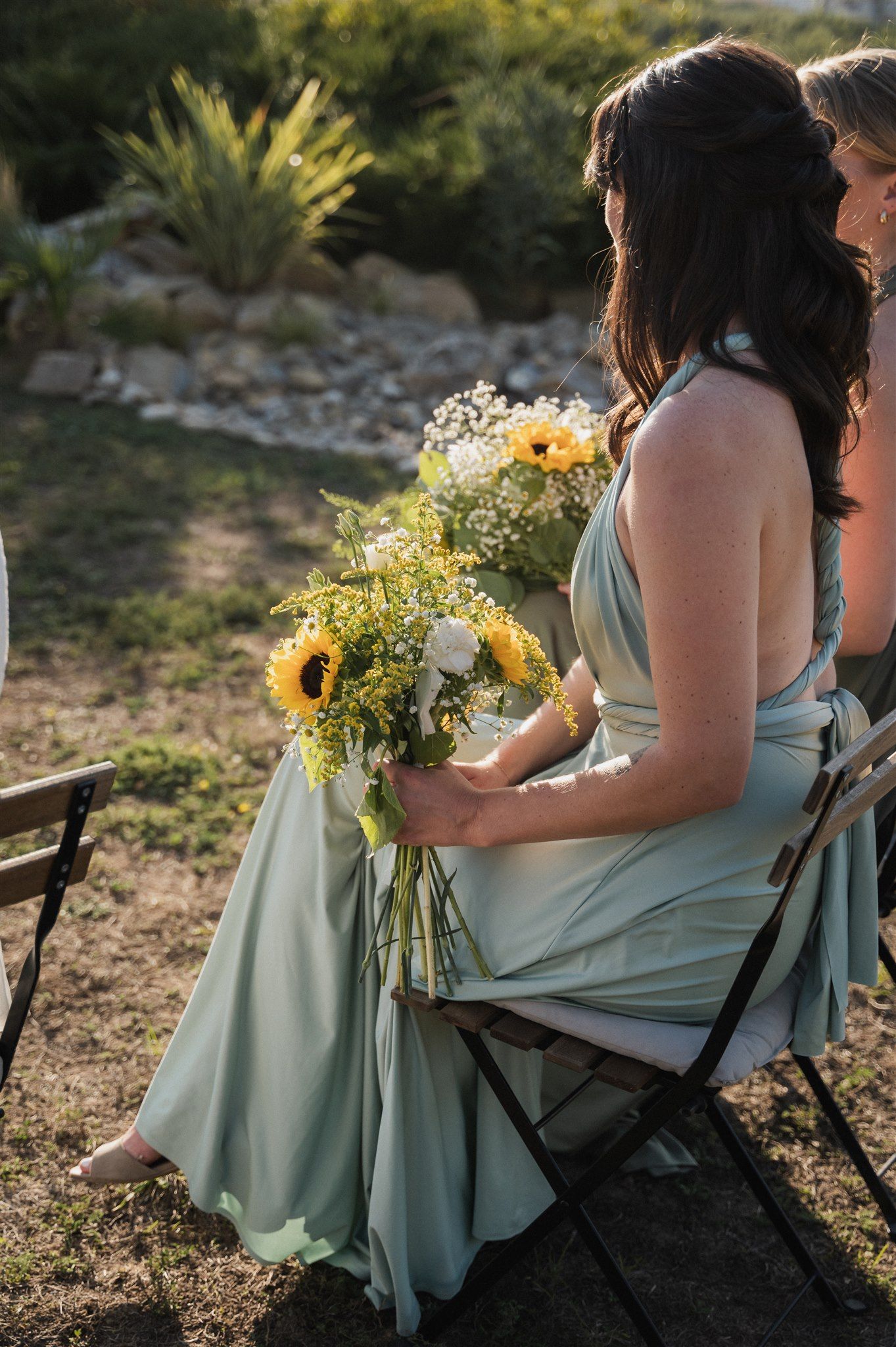Bridesmaids in sage green dresses hold sunflower bouquets at an outdoor event.