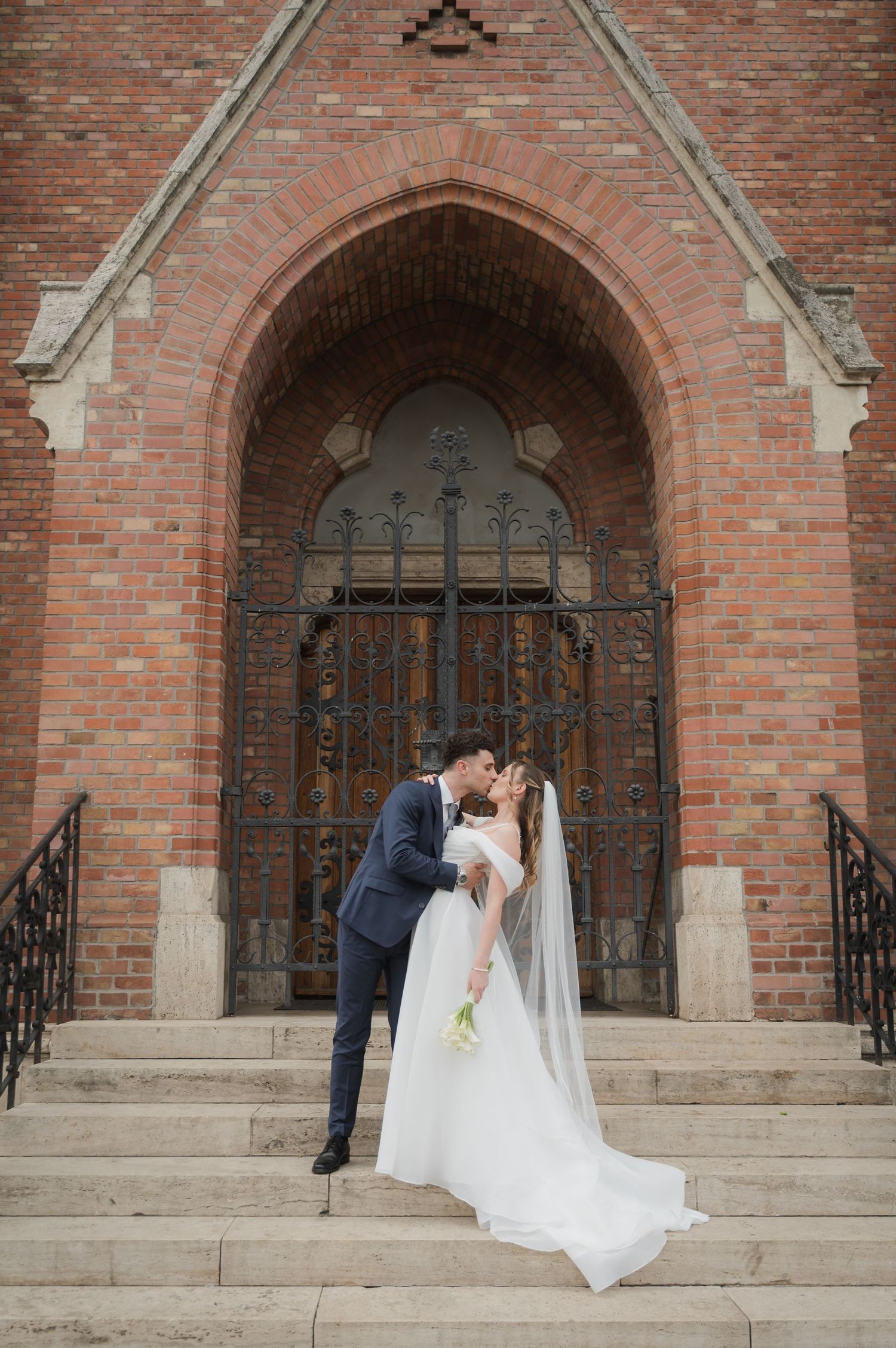 Bride and groom sharing a romantic kiss on church steps beneath a grand Gothic brick archway.