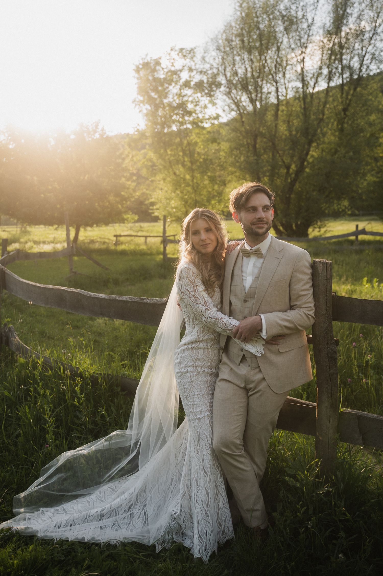 Couple in wedding attire poses together at sunset by a rustic wooden fence surrounded by lush green countryside.