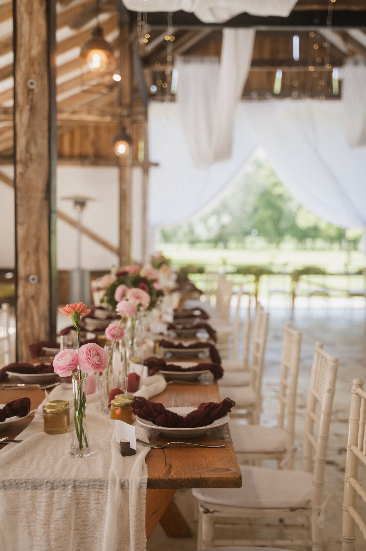 Rustic barn wedding reception table with pink flowers in vases, white linens, and wooden chairs along a long farmhouse table.