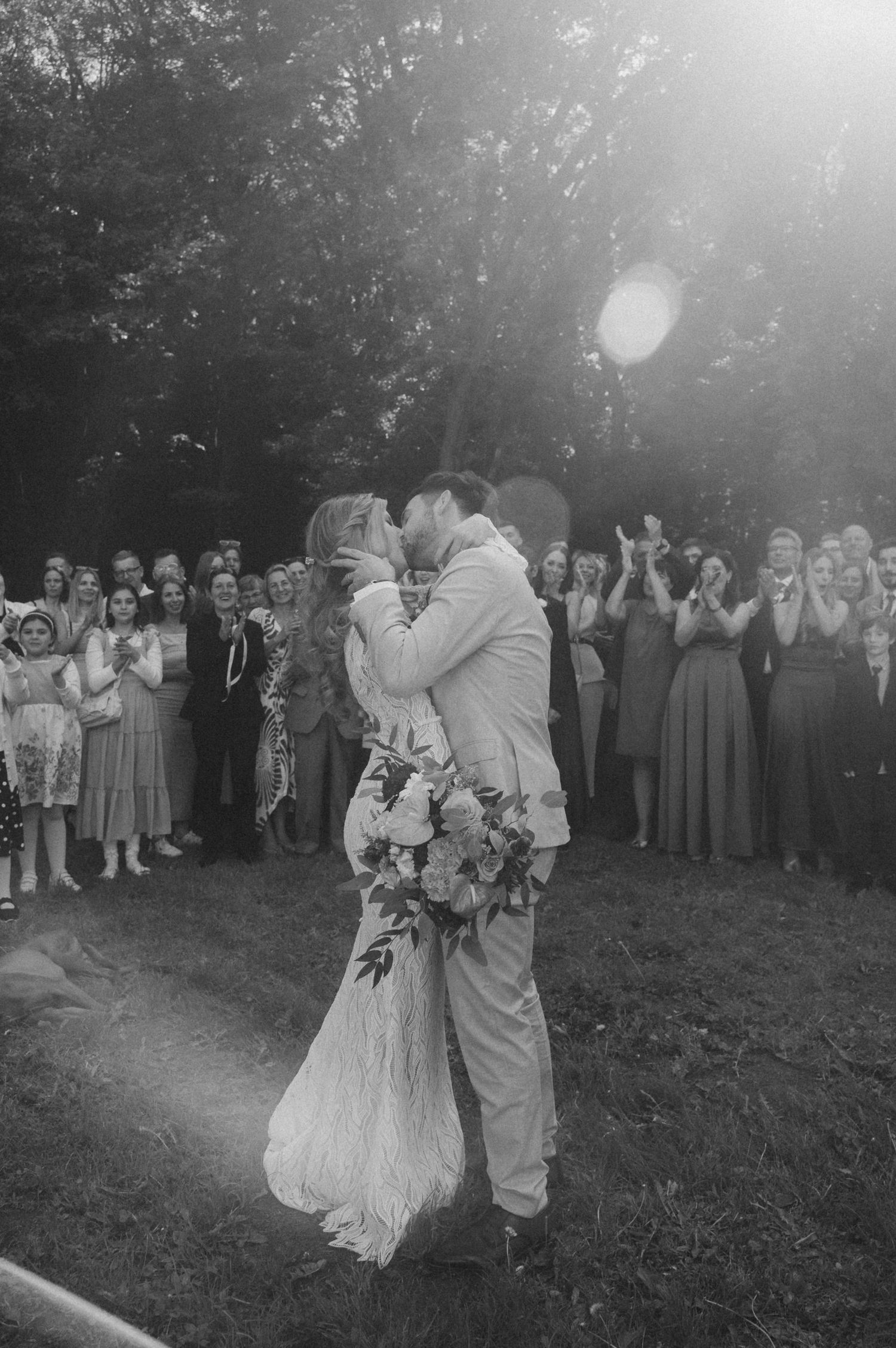 A romantic black and white wedding photo showing a couple's first kiss as guests look on in an outdoor garden setting.
