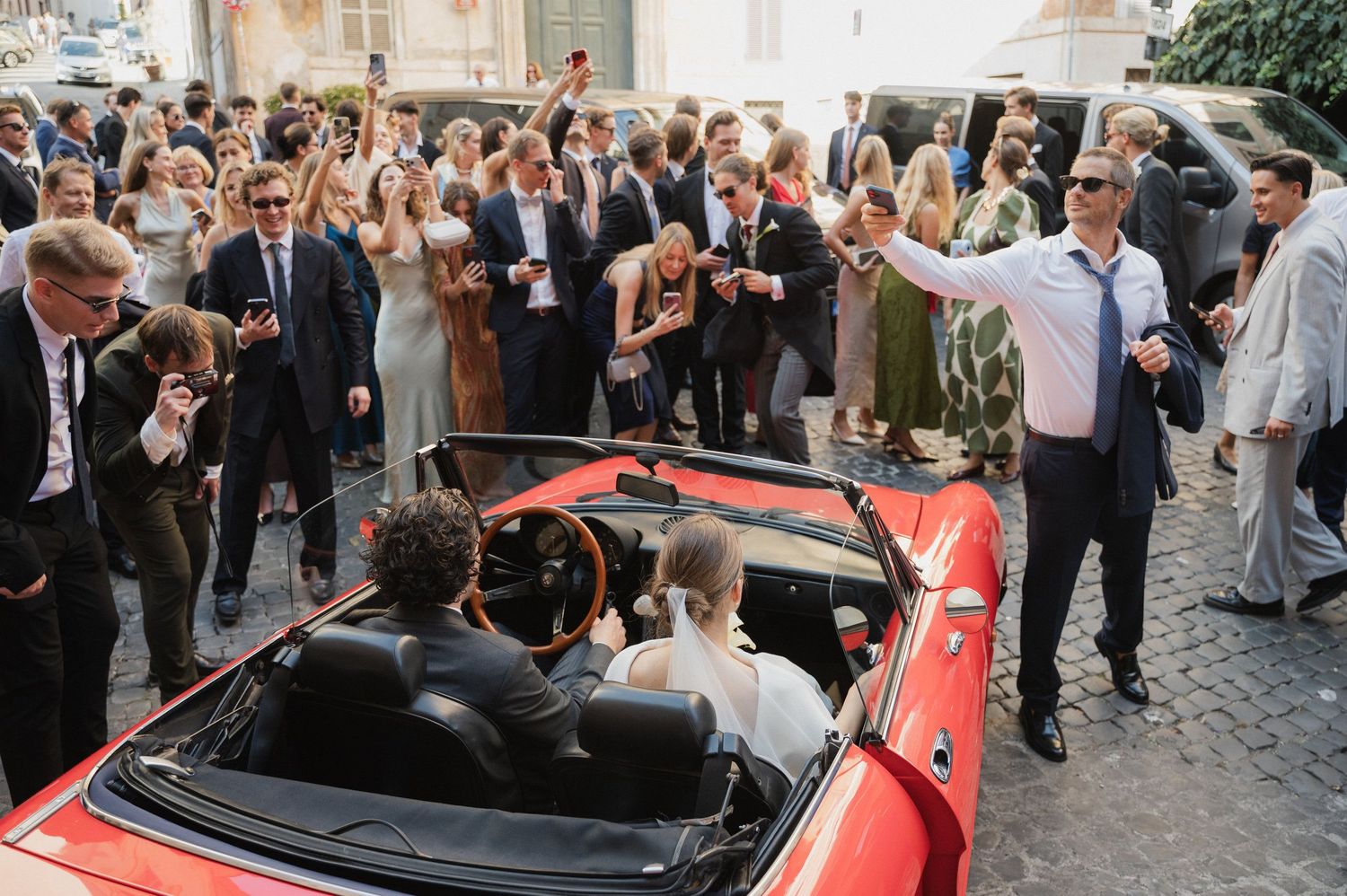 Wedding guests celebrate around a red convertible car on a charming European street.