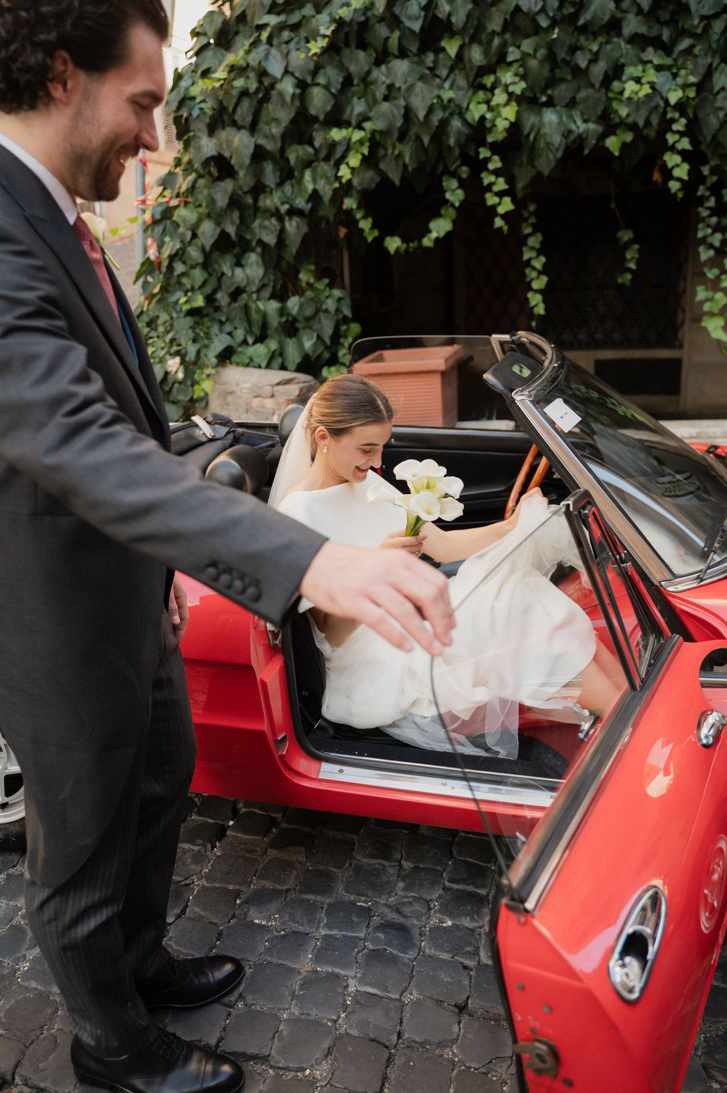 Groom helps bride into classic red convertible car surrounded by ivy-covered walls.