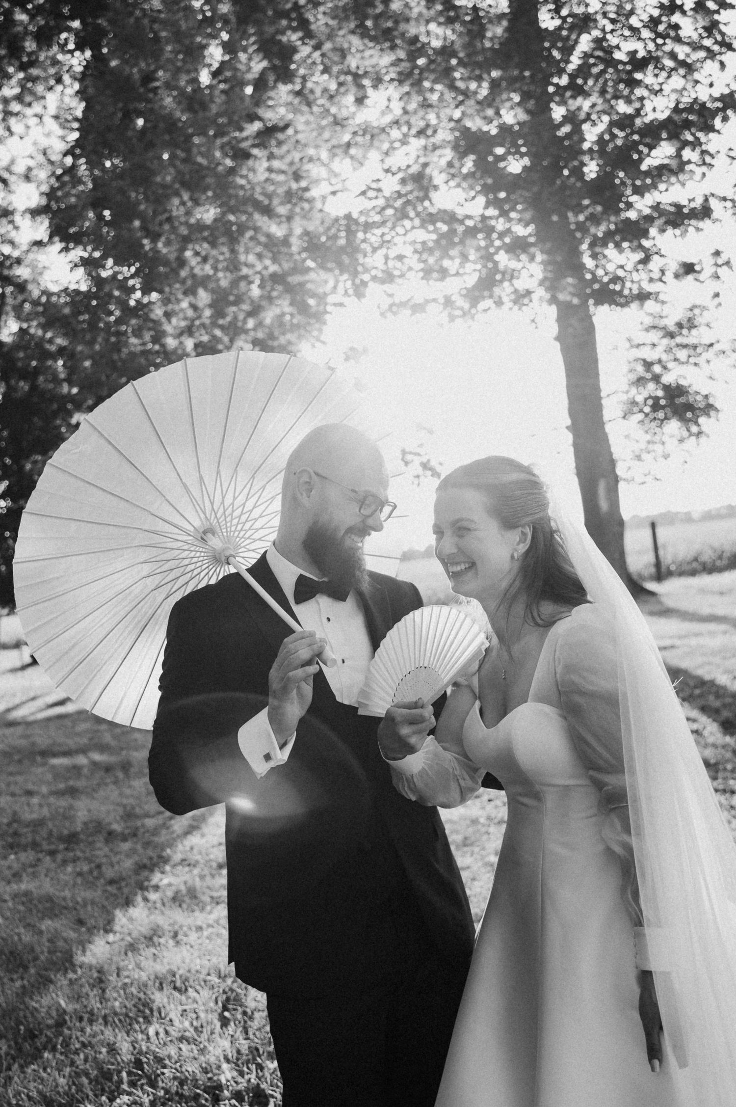 A wedding couple shares an intimate moment while holding white parasols in a sunlit garden setting.