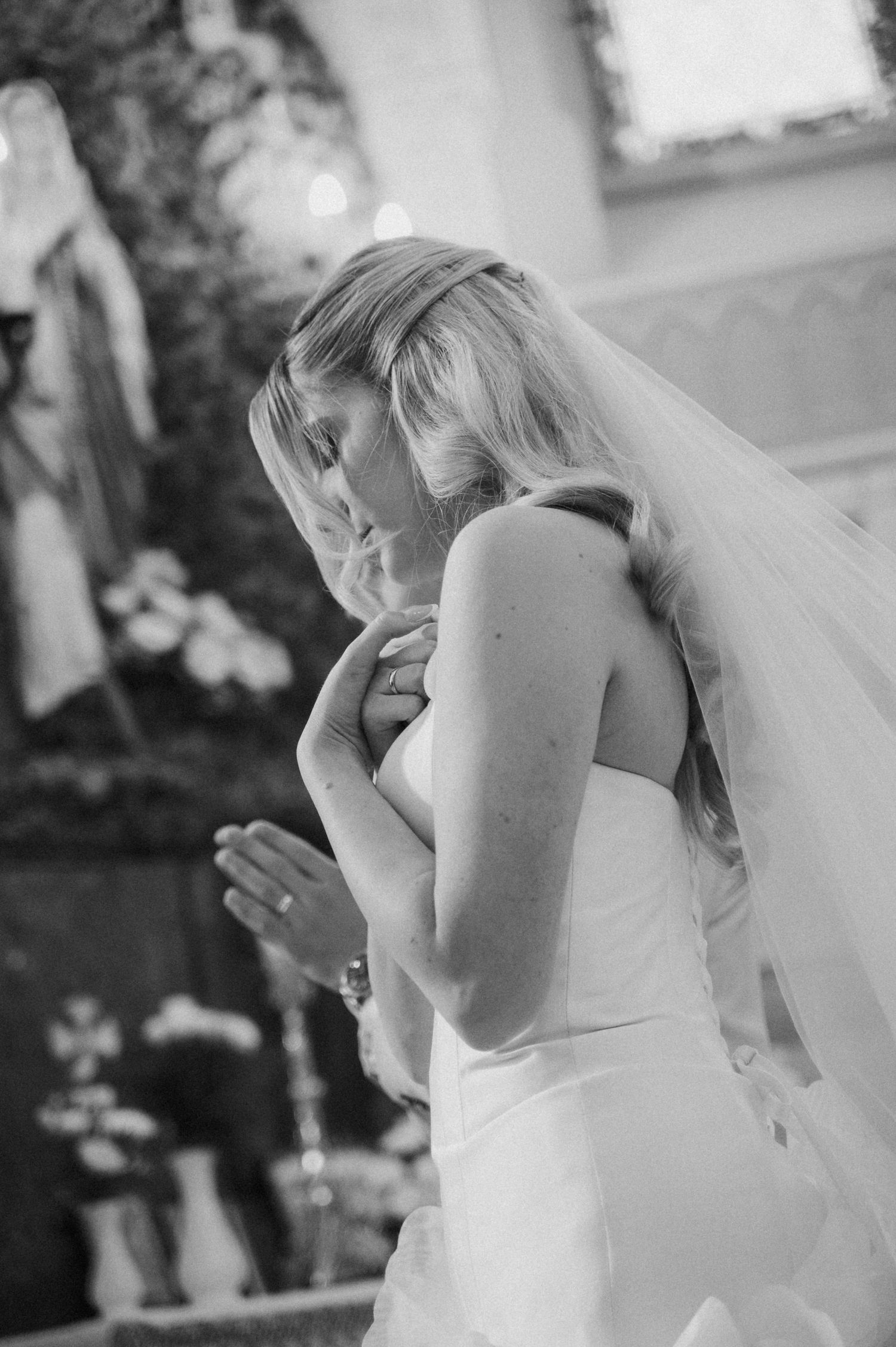 Bride in white strapless gown and veil praying with hands clasped in a church before her wedding ceremony.