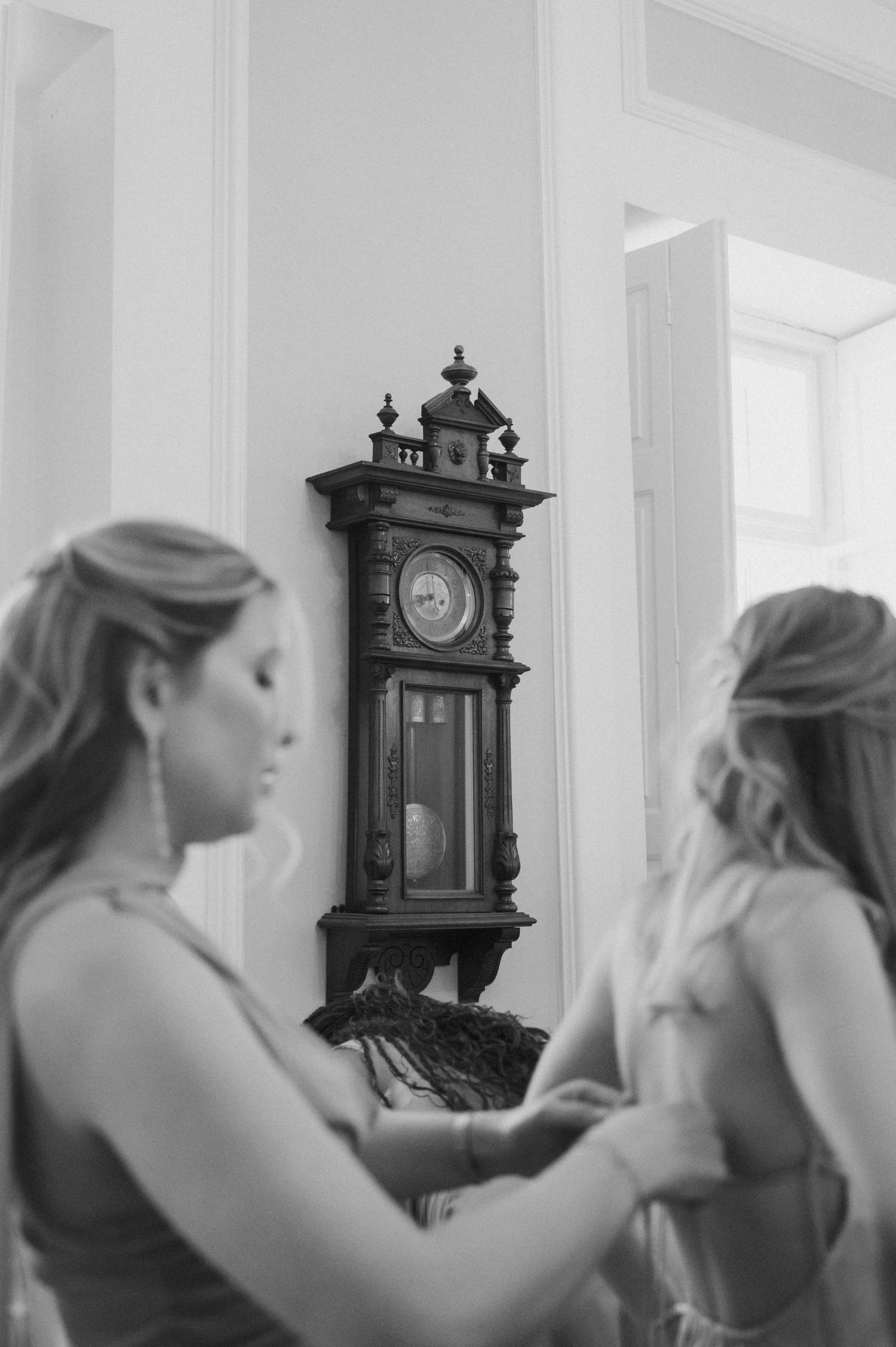 Bridesmaids helping each other get ready in black and white with vintage wall clock in background.