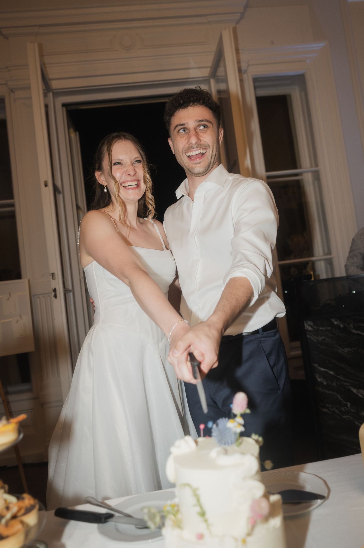 Smiling bride and groom cutting white wedding cake together at elegant indoor reception.
