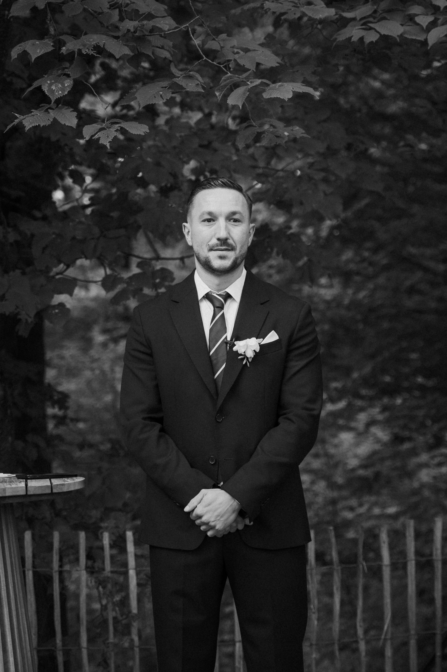 A groom in a black suit with striped tie and boutonniere stands outdoors among lush trees in a black and white photo.
