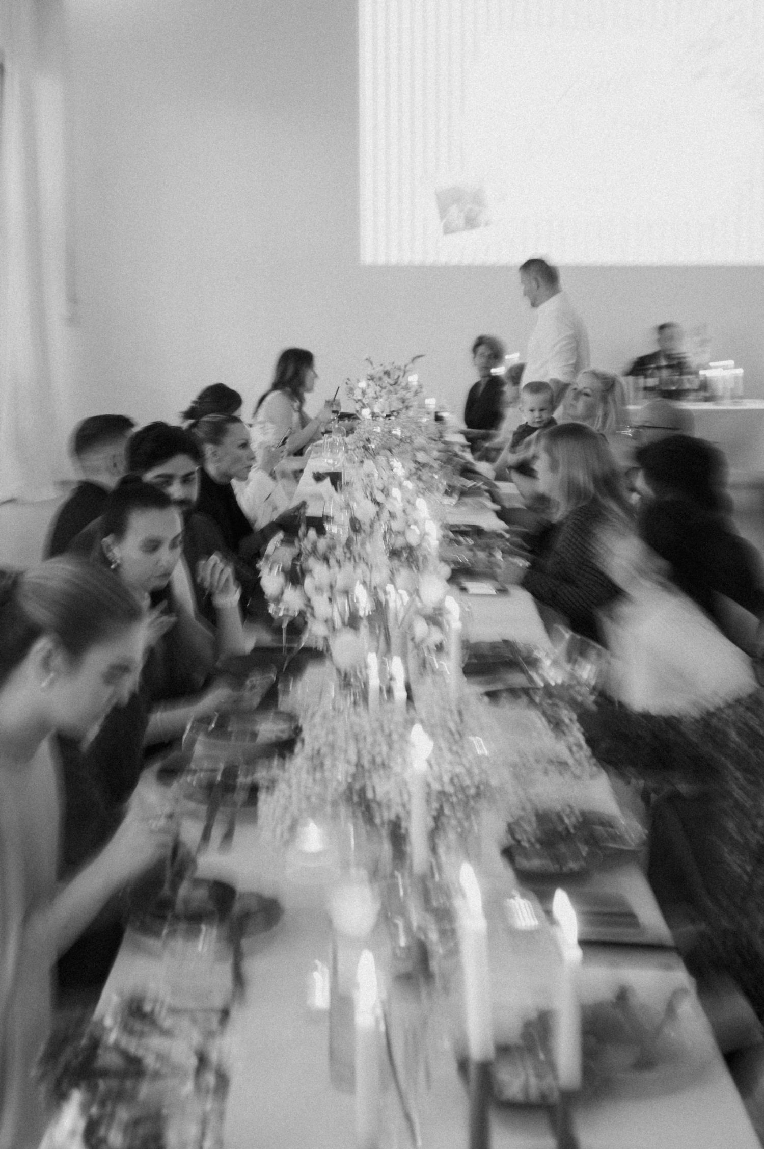 Wedding guests seated at long decorated reception table with floral centerpieces and candles in black and white.