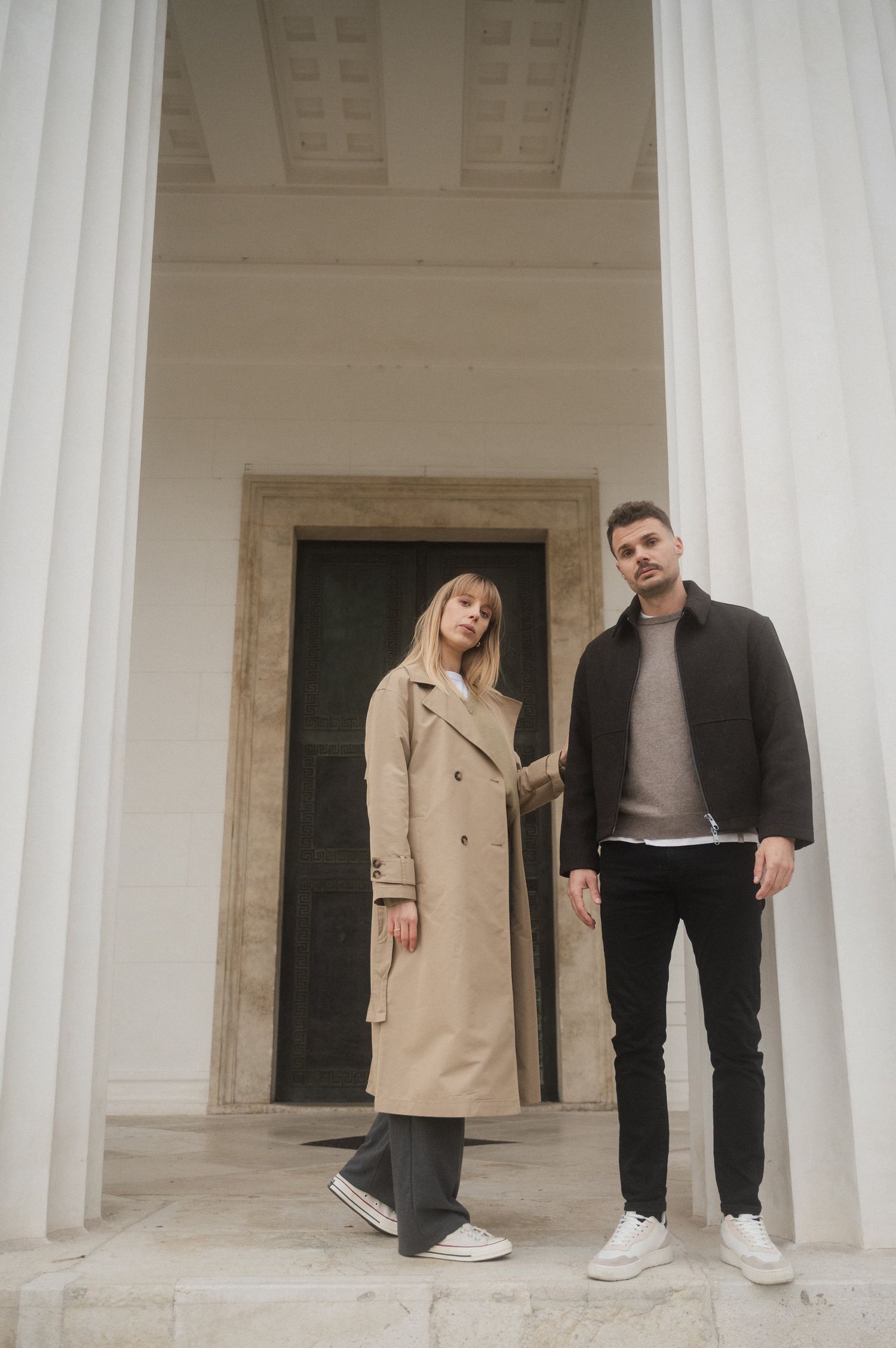 A couple in casual attire poses between white columns in a modern architectural setting.