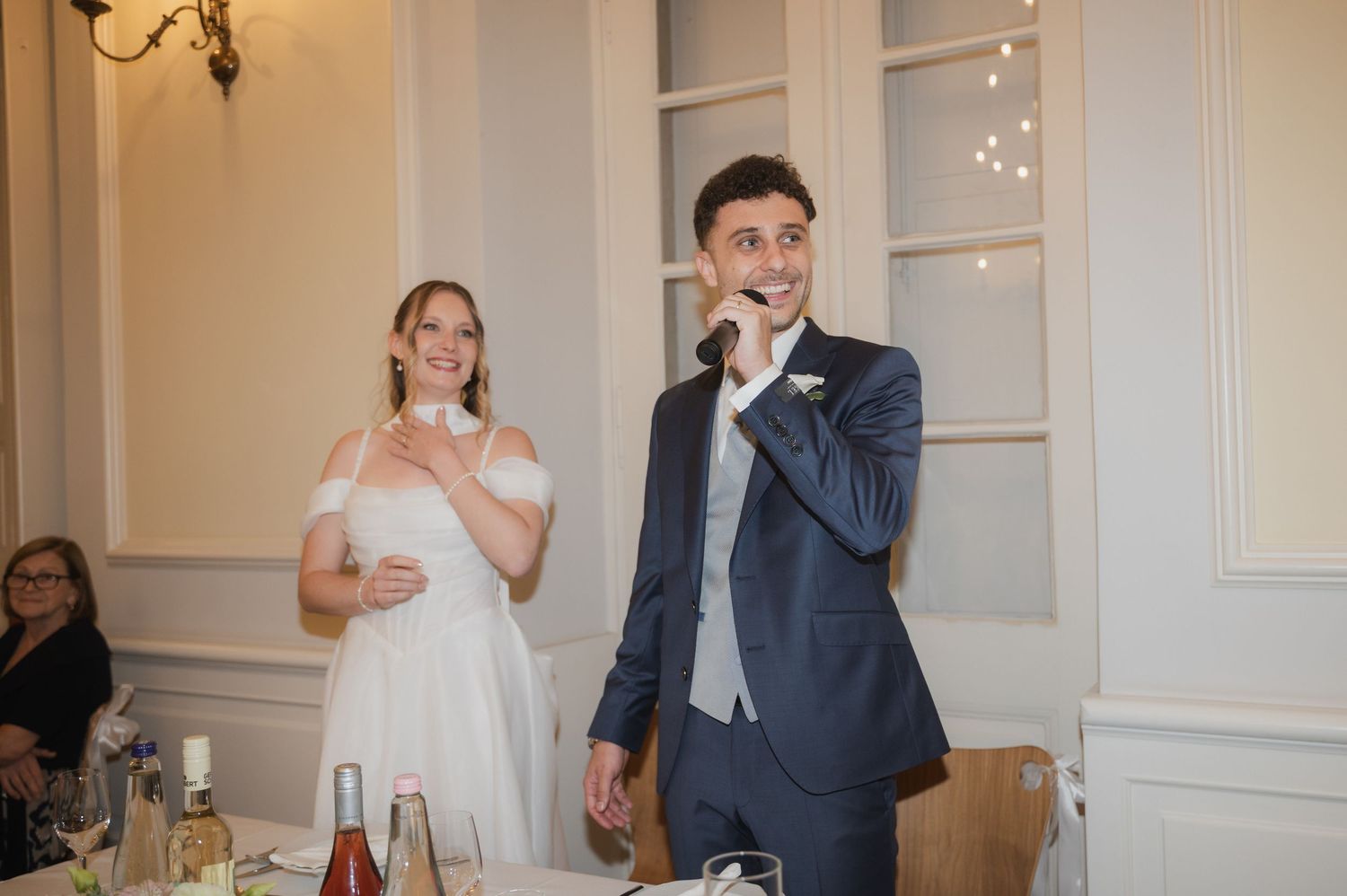Groom speaks into a microphone at wedding reception while smiling bride stands beside him.
