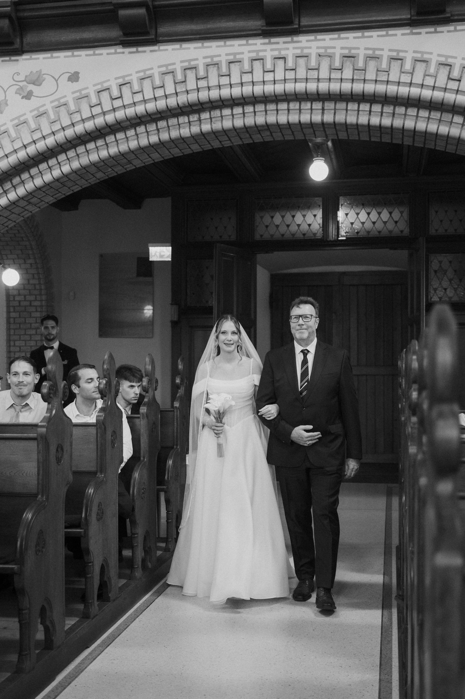 Black and white photo of a bride walking down the church aisle with her escort during a wedding ceremony.
