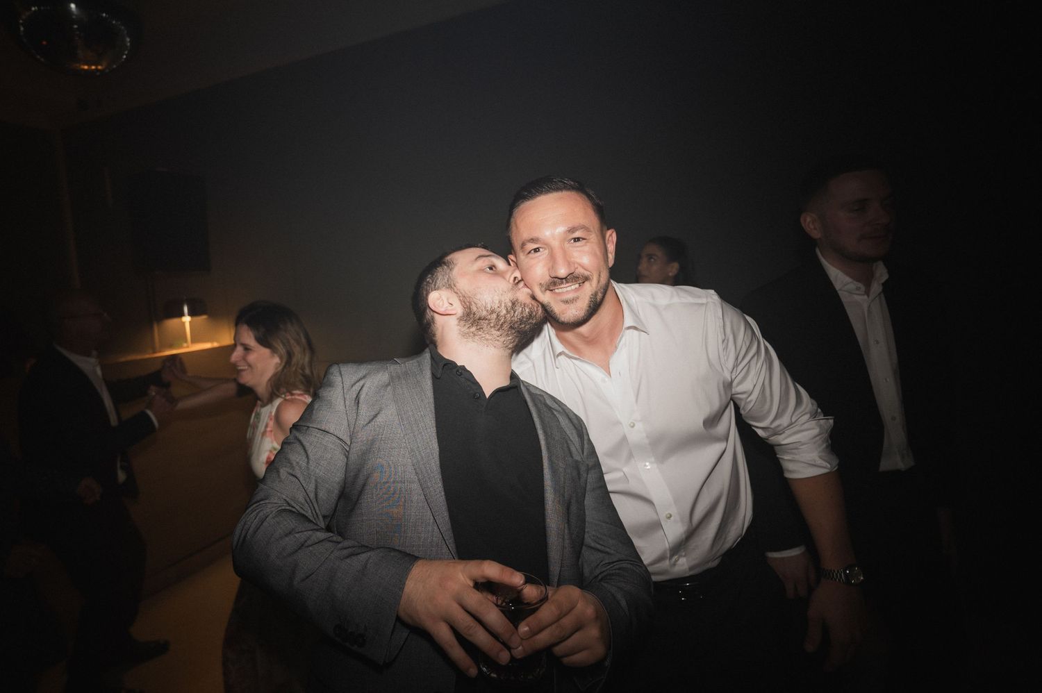 Two men laughing and sharing a kiss on the cheek at a lively wedding party.