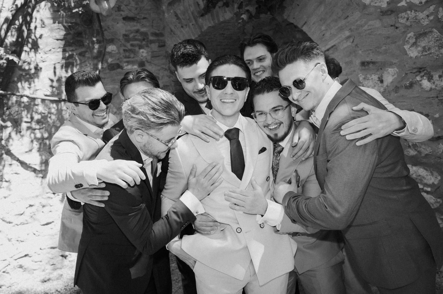 Groom surrounded by groomsmen in sunglasses sharing a joyful group hug in black and white photo.