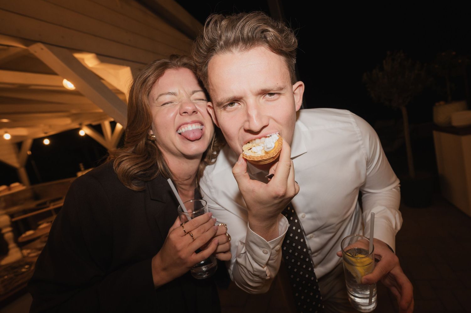 Two friends laugh and make silly faces while enjoying cupcakes at an outdoor evening event.
