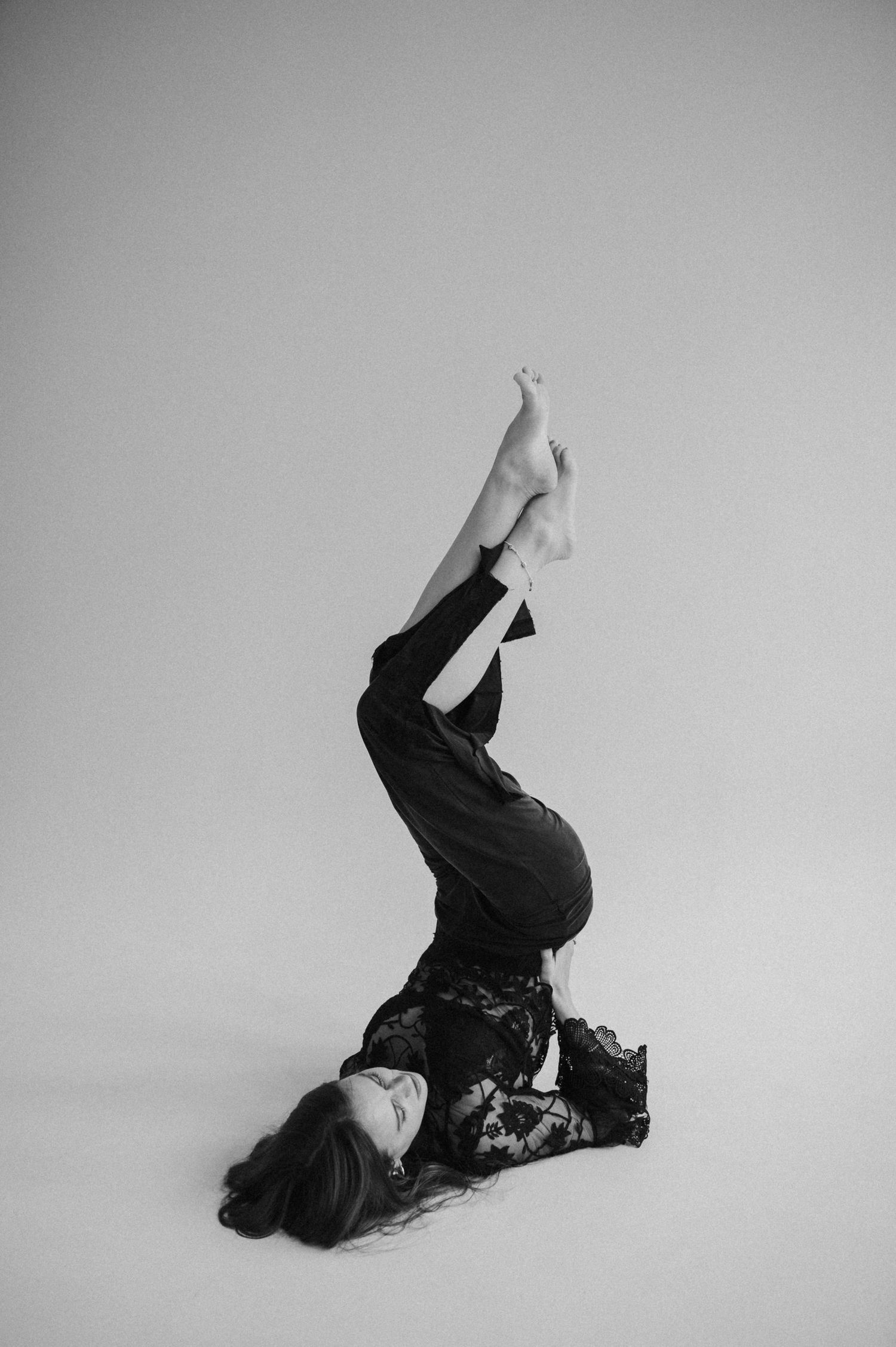 A person in black clothing lies on a white floor with legs raised in an artistic pose against a light background.