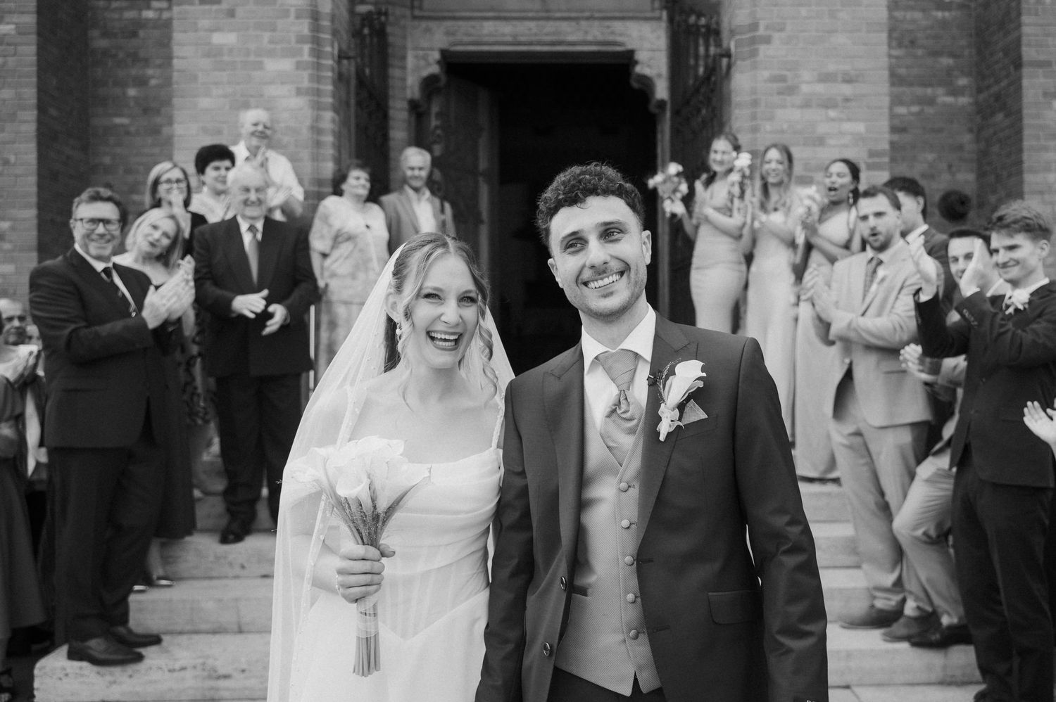 Black and white photo of a smiling bride and groom walking out of a church, surrounded by cheering wedding guests.