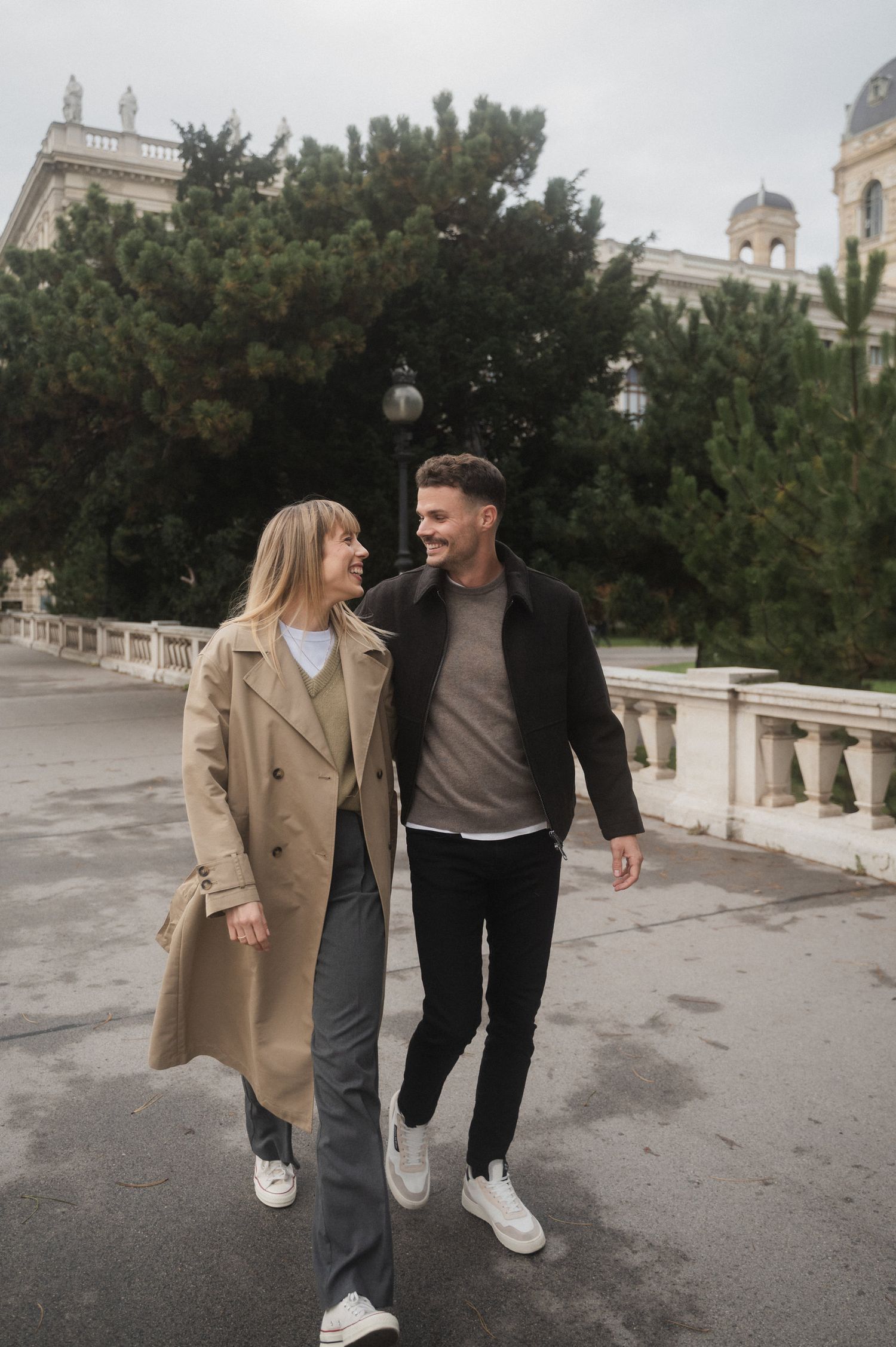 A couple in casual attire walks together on a paved pathway near ornate architecture on a cloudy day.