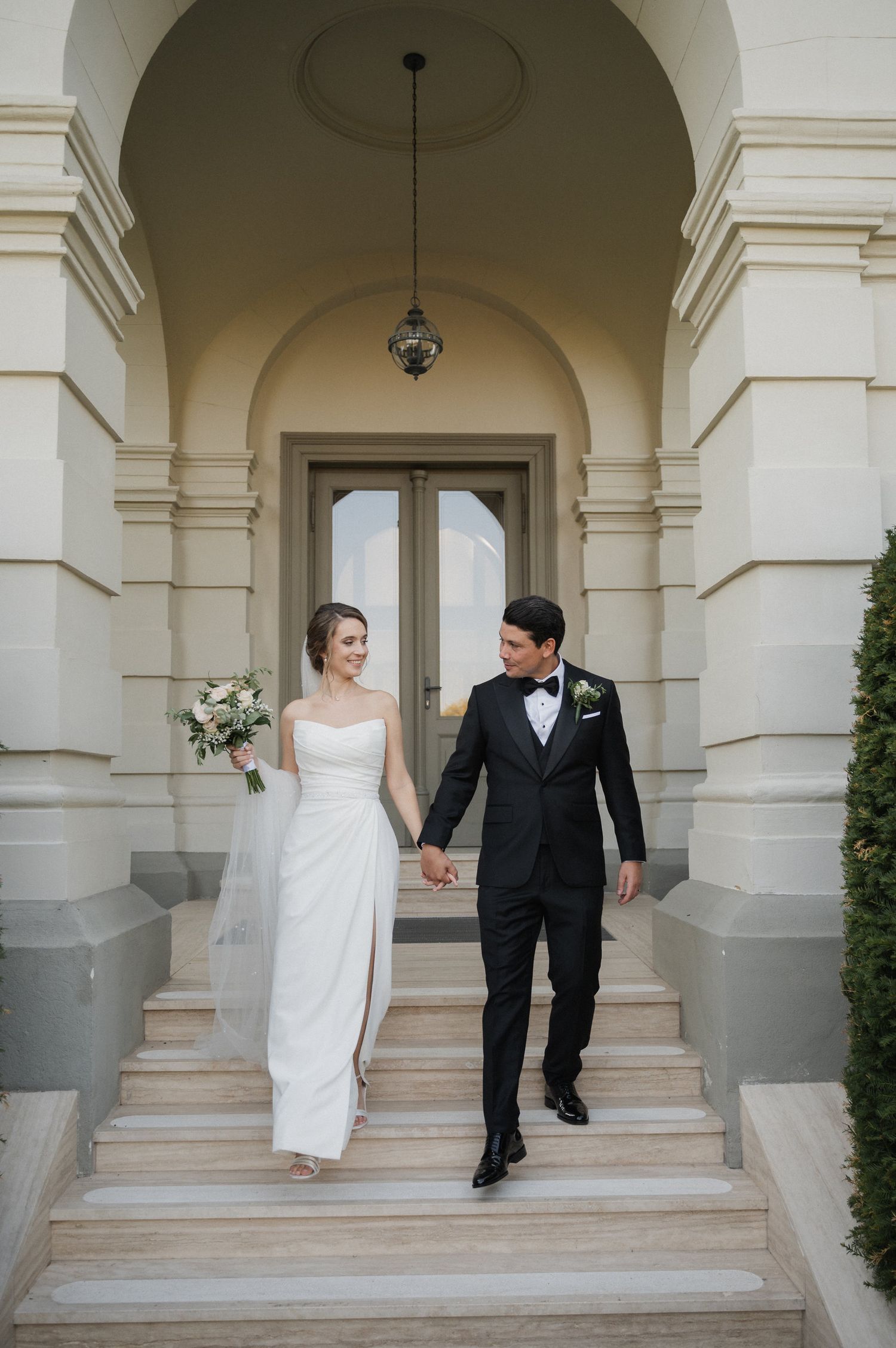 A couple in formal wedding attire walks through elegant white archways in a classical architectural setting.