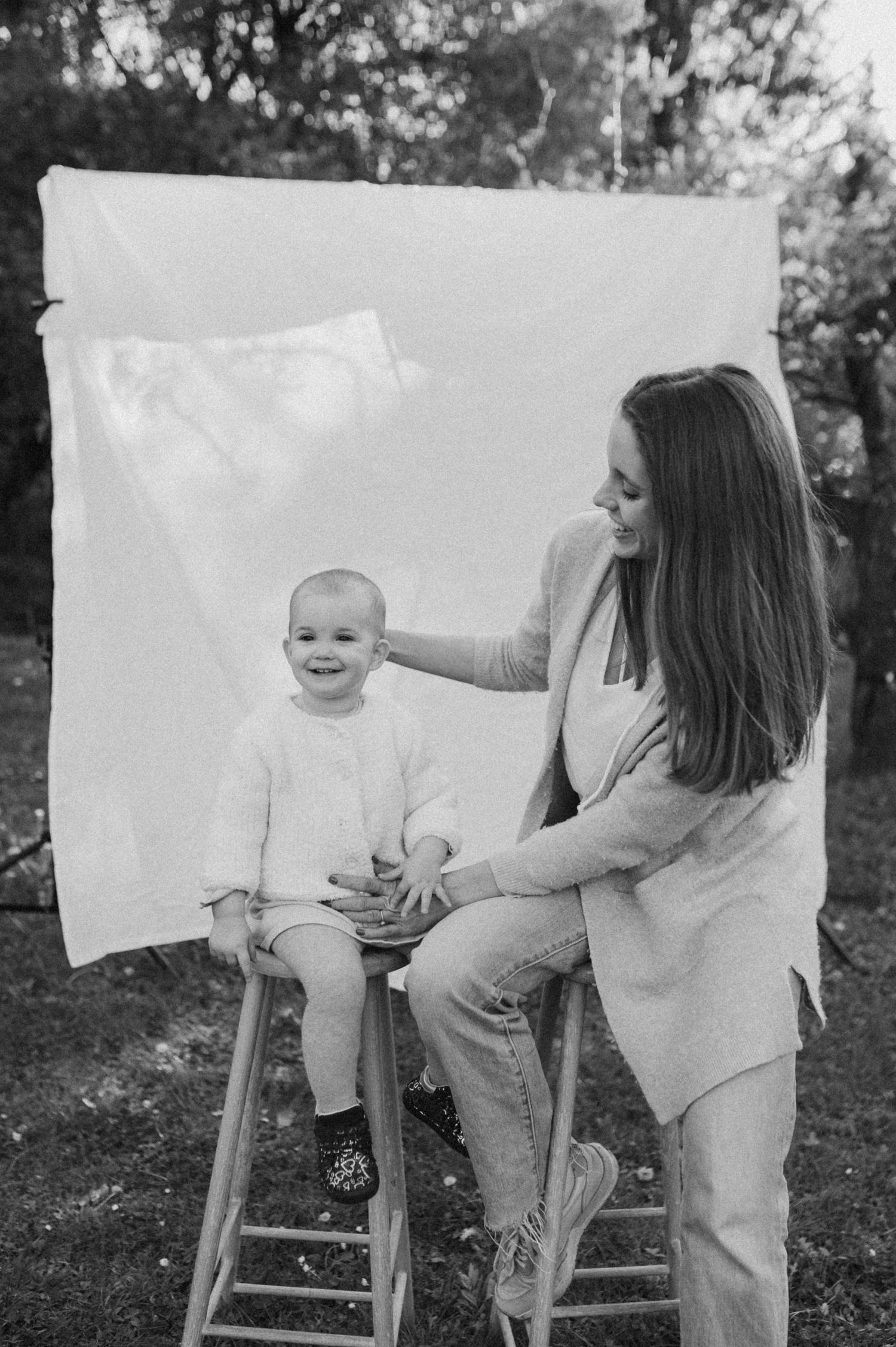 Black and white photo of a smiling baby on a stool with a woman beside them, outdoors with a white backdrop.