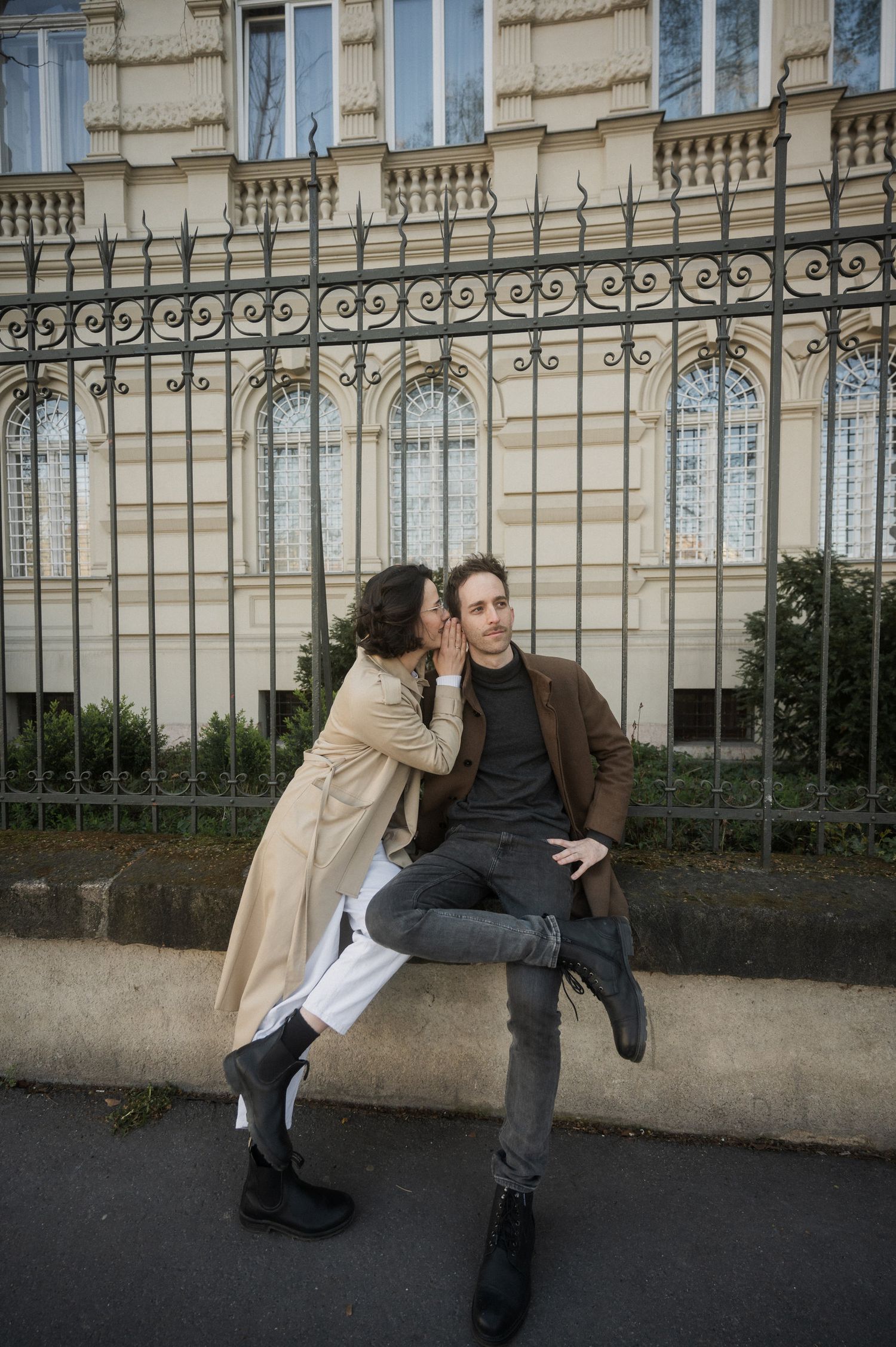 Two people share an intimate moment against an ornate iron fence and classical building facade in Paris.