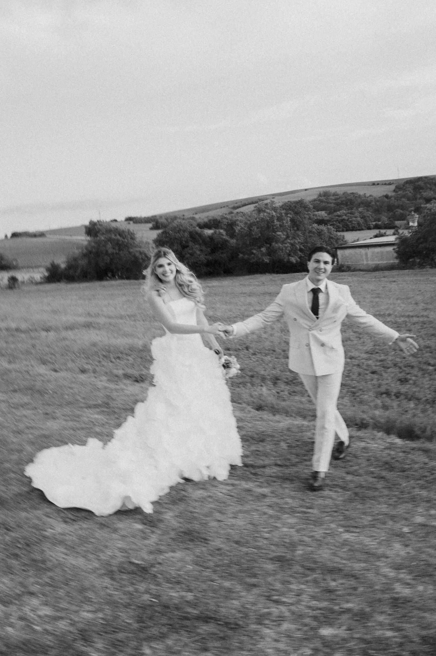 Black and white photo of bride and groom holding hands running through an open countryside field.