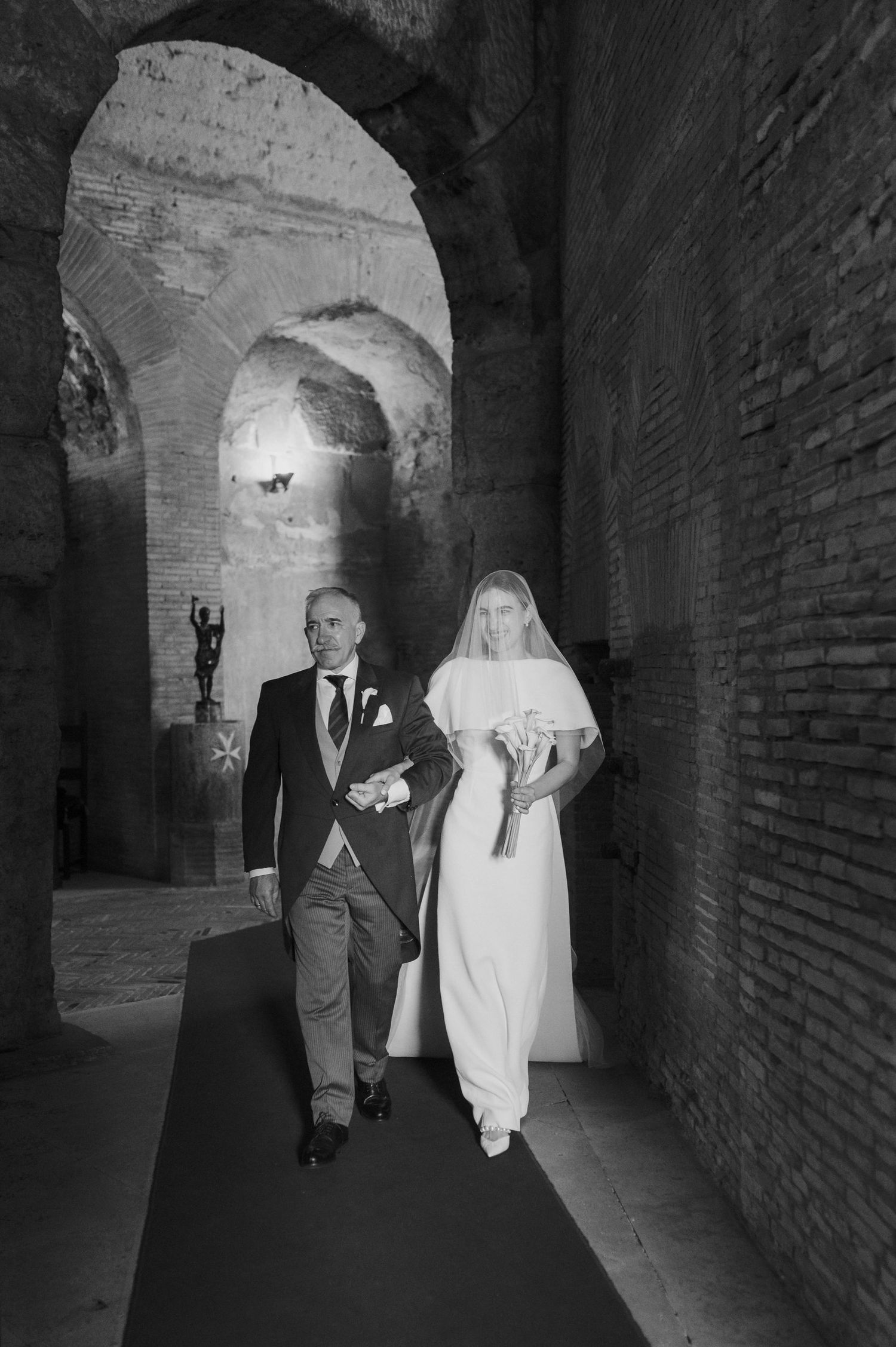 Wedding party walking down a dark stone corridor with dramatic arched lighting.