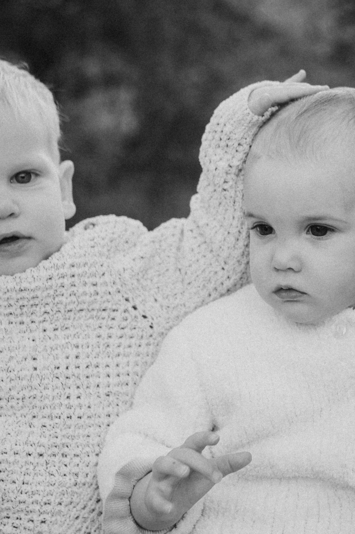 Black and white photo of two babies in knit sweaters, one resting hand on the others head.