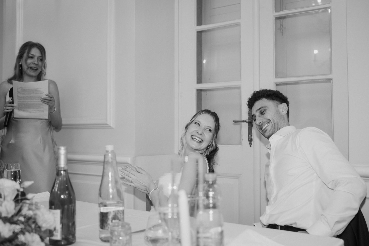 Bride and groom laugh joyfully at wedding reception table as guest delivers speech in background.