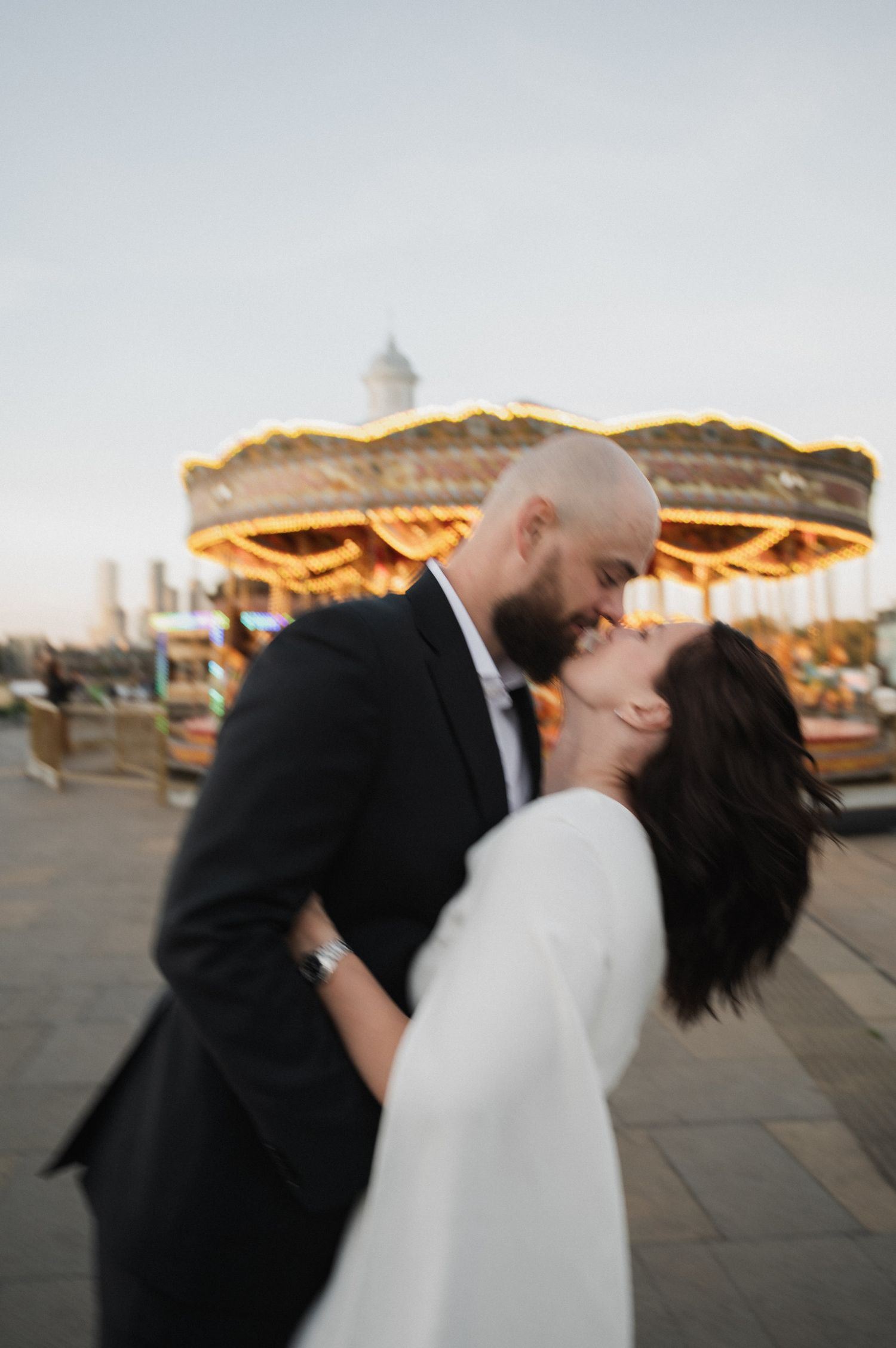 Couple shares intimate moments at sunset with carnival lights in the background.