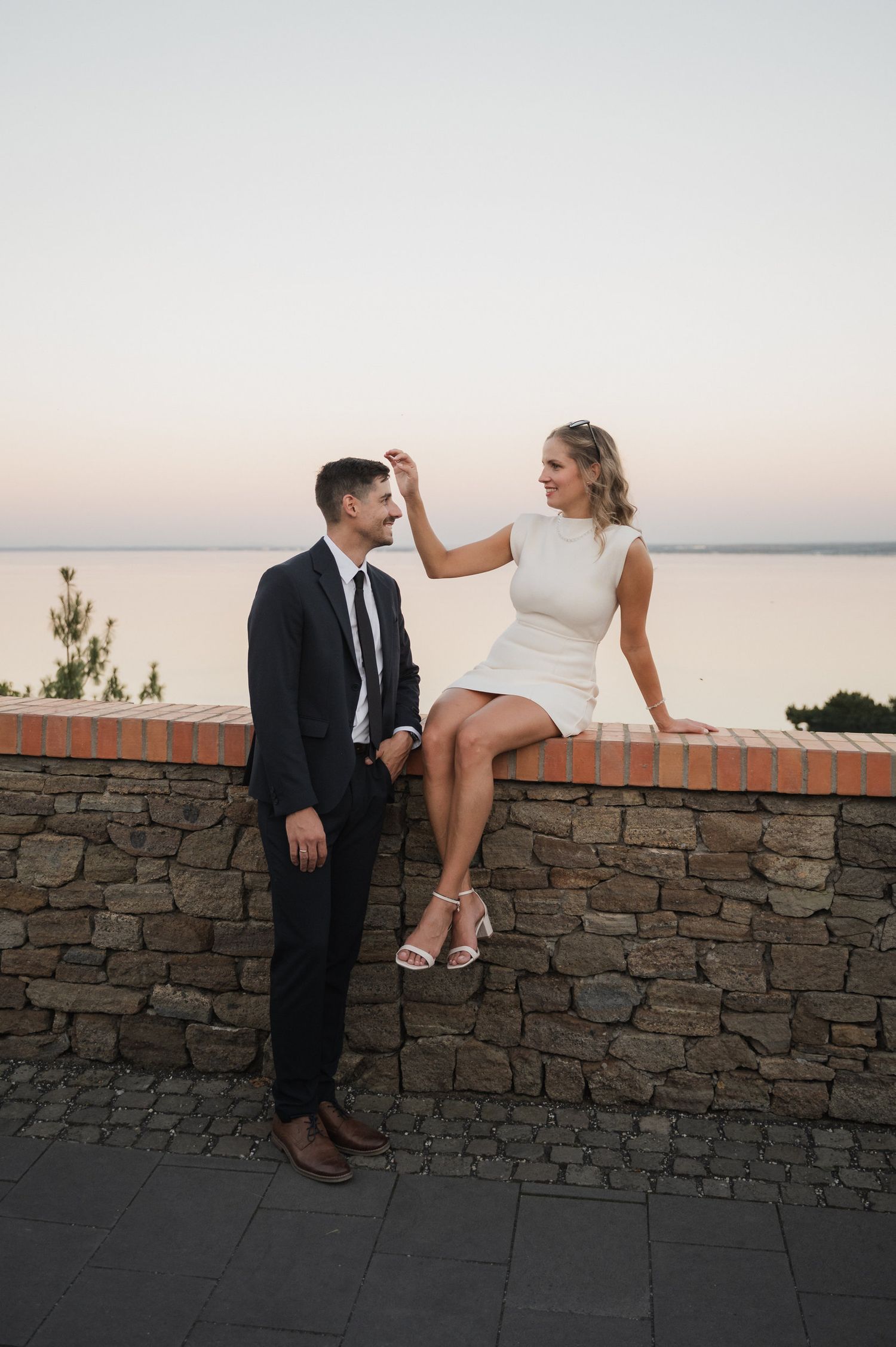 A couple shares a playful moment while sitting on a stone wall during sunset with water views in the distance.