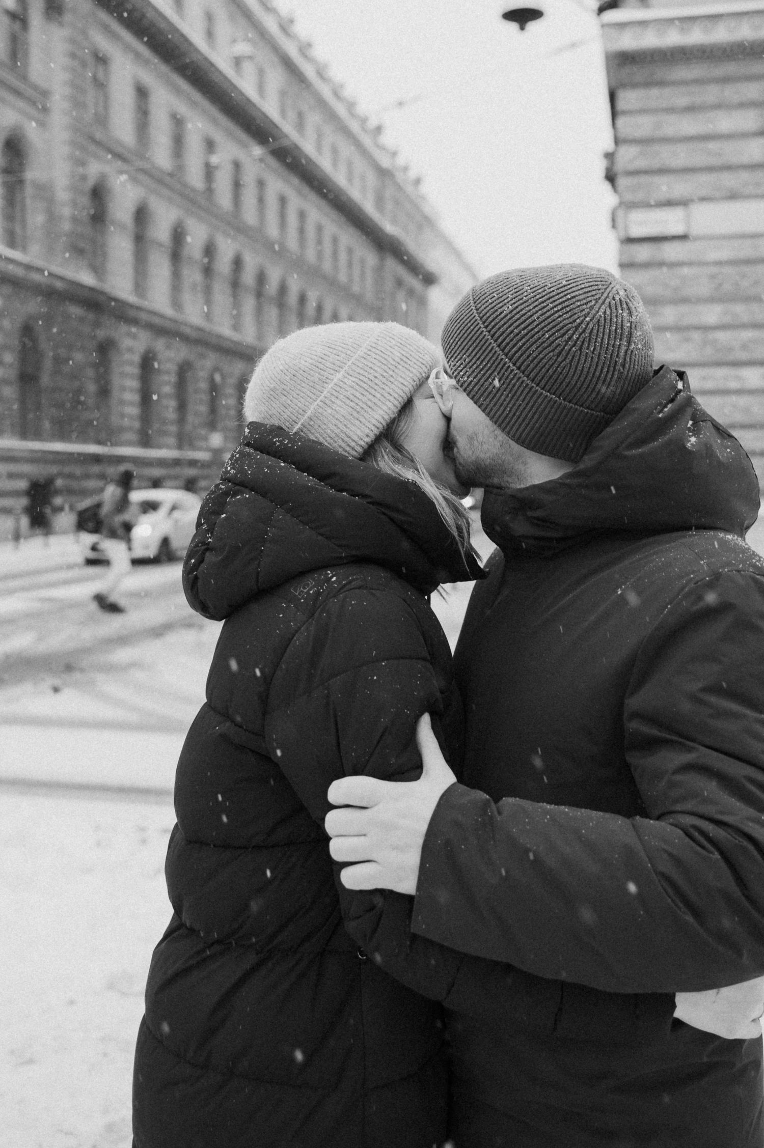 Two people in winter coats and beanies embracing on a snowy day.